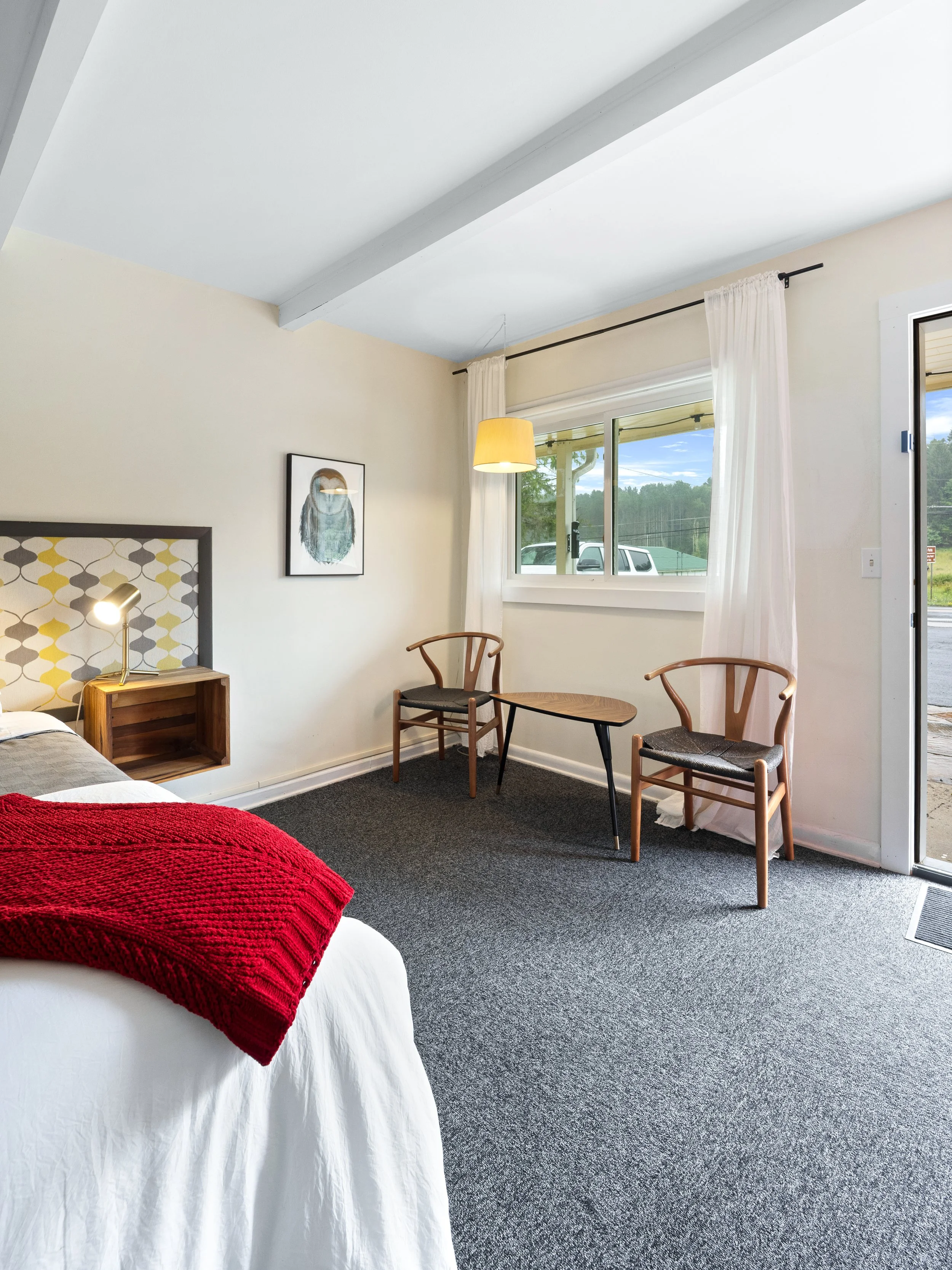 A bedroom with a bed, a nightstand, two chairs, a window with curtains, and a piece of artwork on the wall.