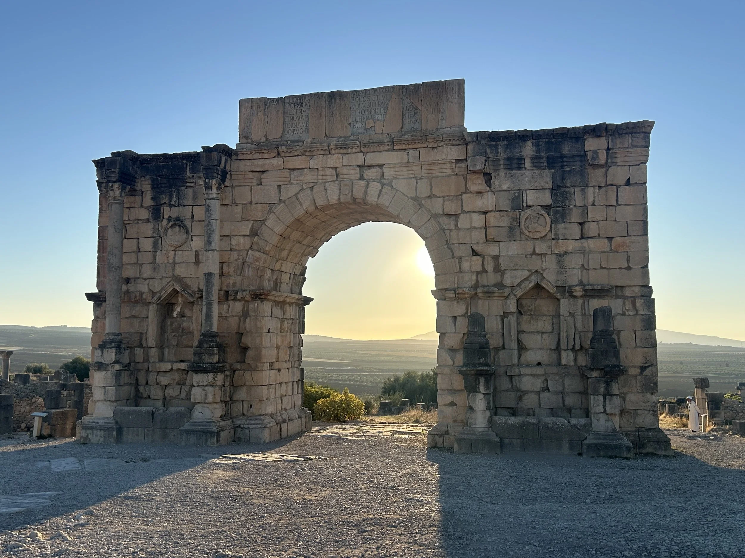 An archway stands alone and has the golden hour sunlight seeping through. Fields stretch through the gateway