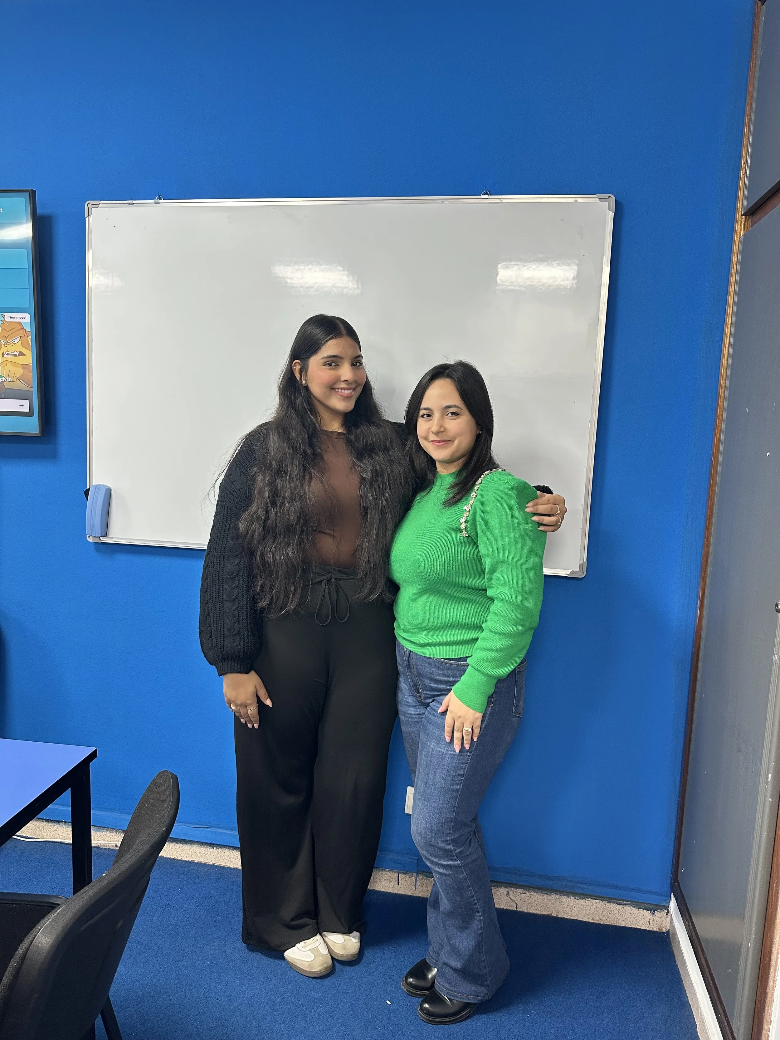 Two young women stand side by side smiling in front of a whiteboard in a classroom