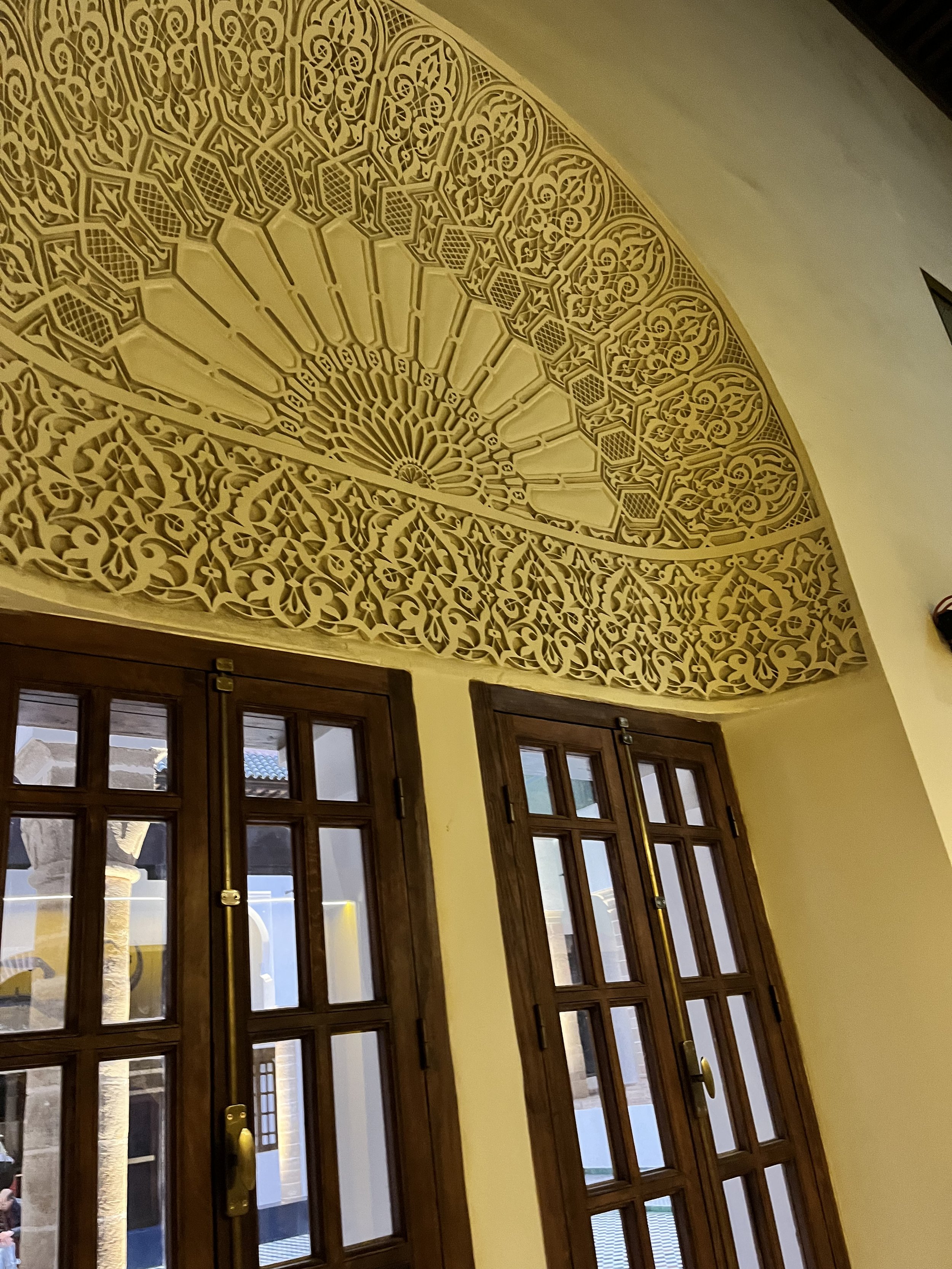 Above two sets of narrow wooden French doors and carved into the wall itself is a shallow bowled half-circle with geometric and floral arabesque designs