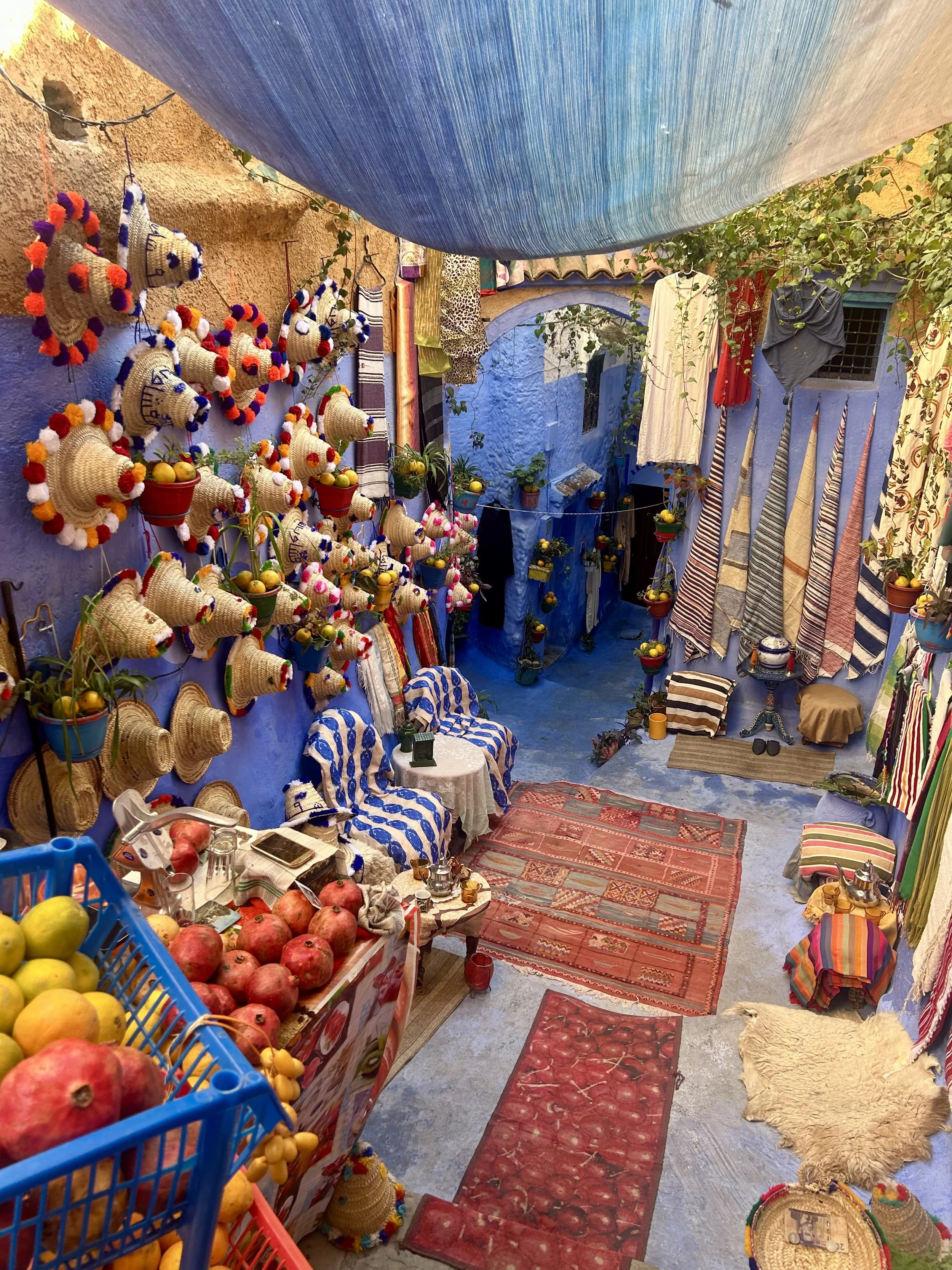 A colorful street showcases Chefchaouen's many wares such as distinct sunhats with pompoms, rugs, blankets, and pomegranates all against the blue walls of the city