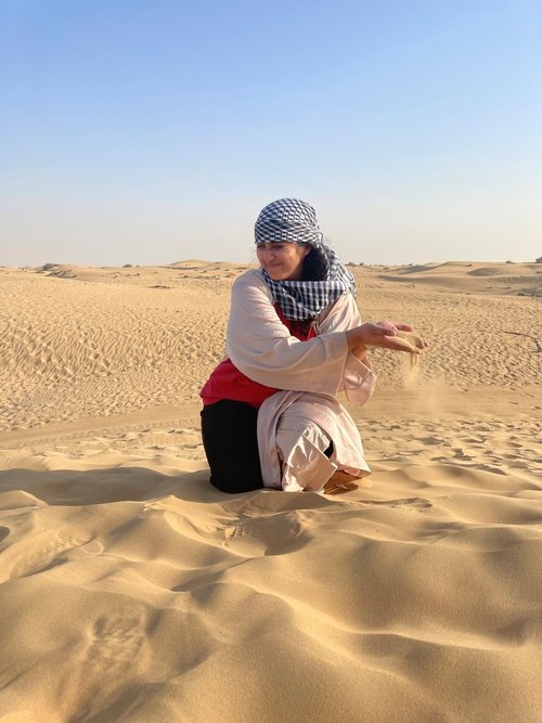 Hajrah kneels in the sand, which stretches into the distance. She wears a blue checkered scarf around her head