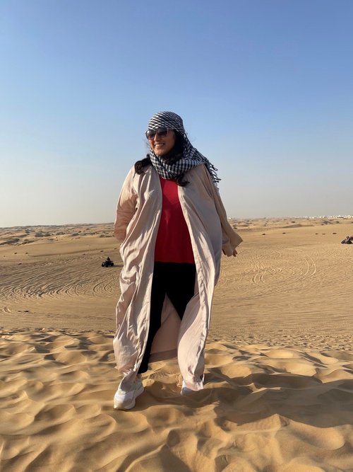 Hajrah stands in the sand, smiling. She's wearing a long flowing coat and a blue checkered scarf around her head.