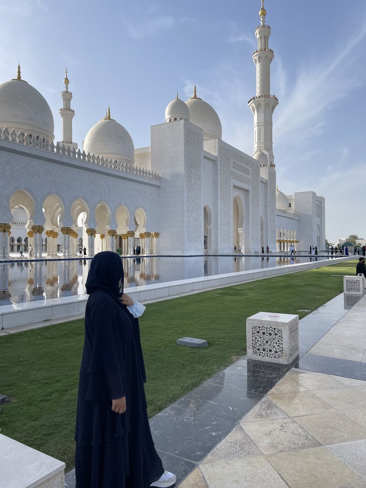 A woman in a black abaya and headscarf stands with her back to the camera looking at a large, new white mosque with gold detailing.