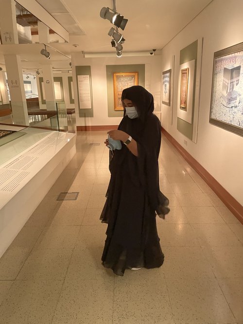 A woman in a black abaya and scarf looks at a camera in her hands. She stands in a museum gallery with photos of Islamic religious sites hanging on the walls