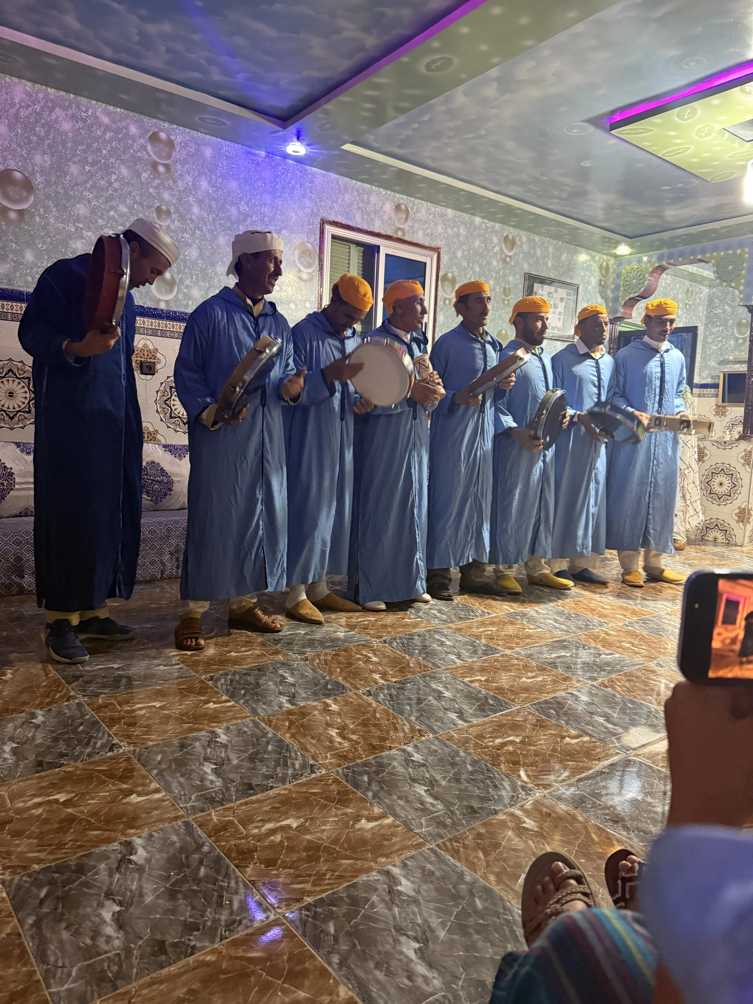 8 men in light blow robes and either white or orange caps stand in a line playing large tambourines in a hall.