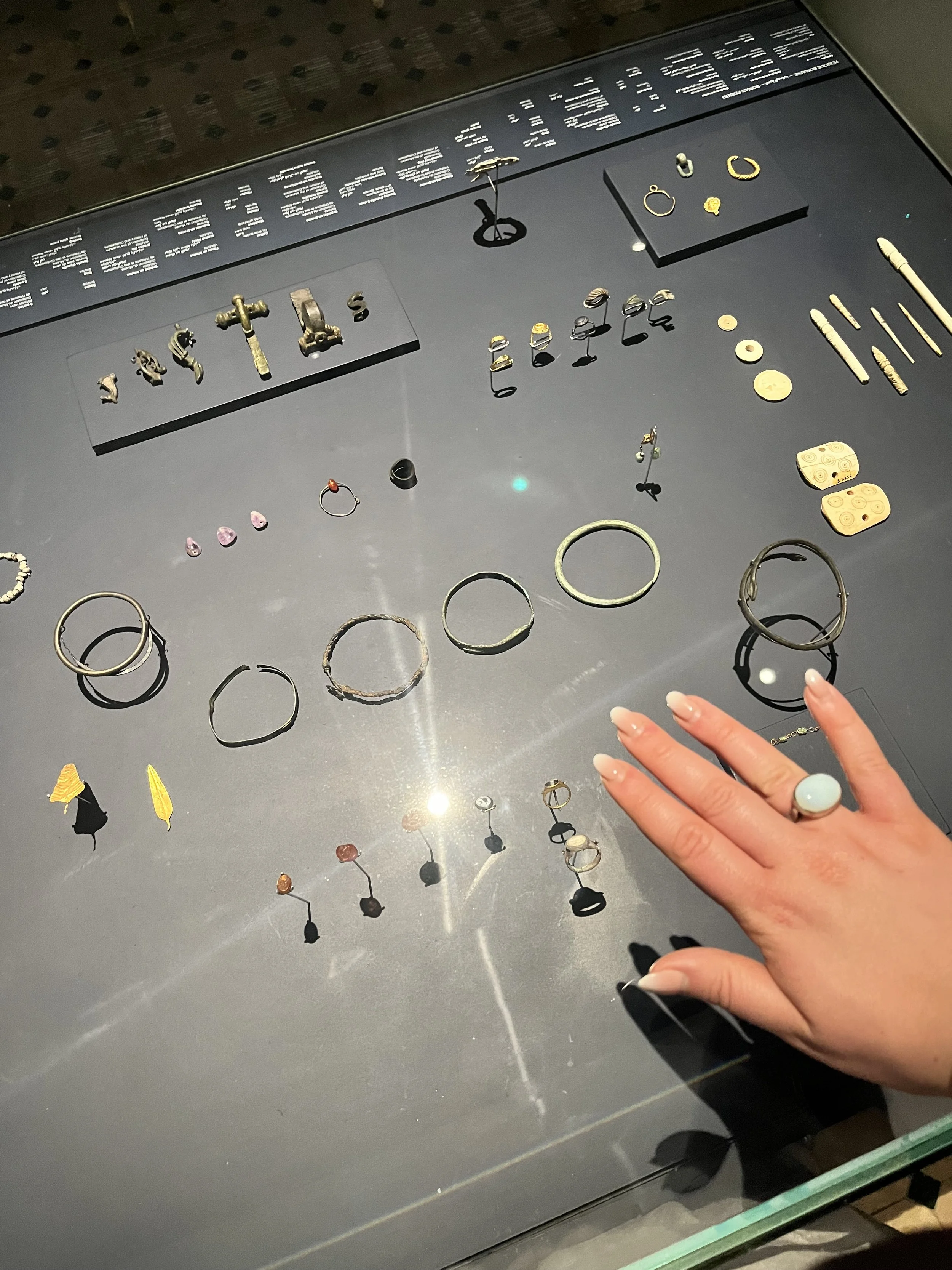 A hand rests on the glass top of a museum display case. Inside the case are ancient jewelry pieces like earrings, bangles, pins, and rings.