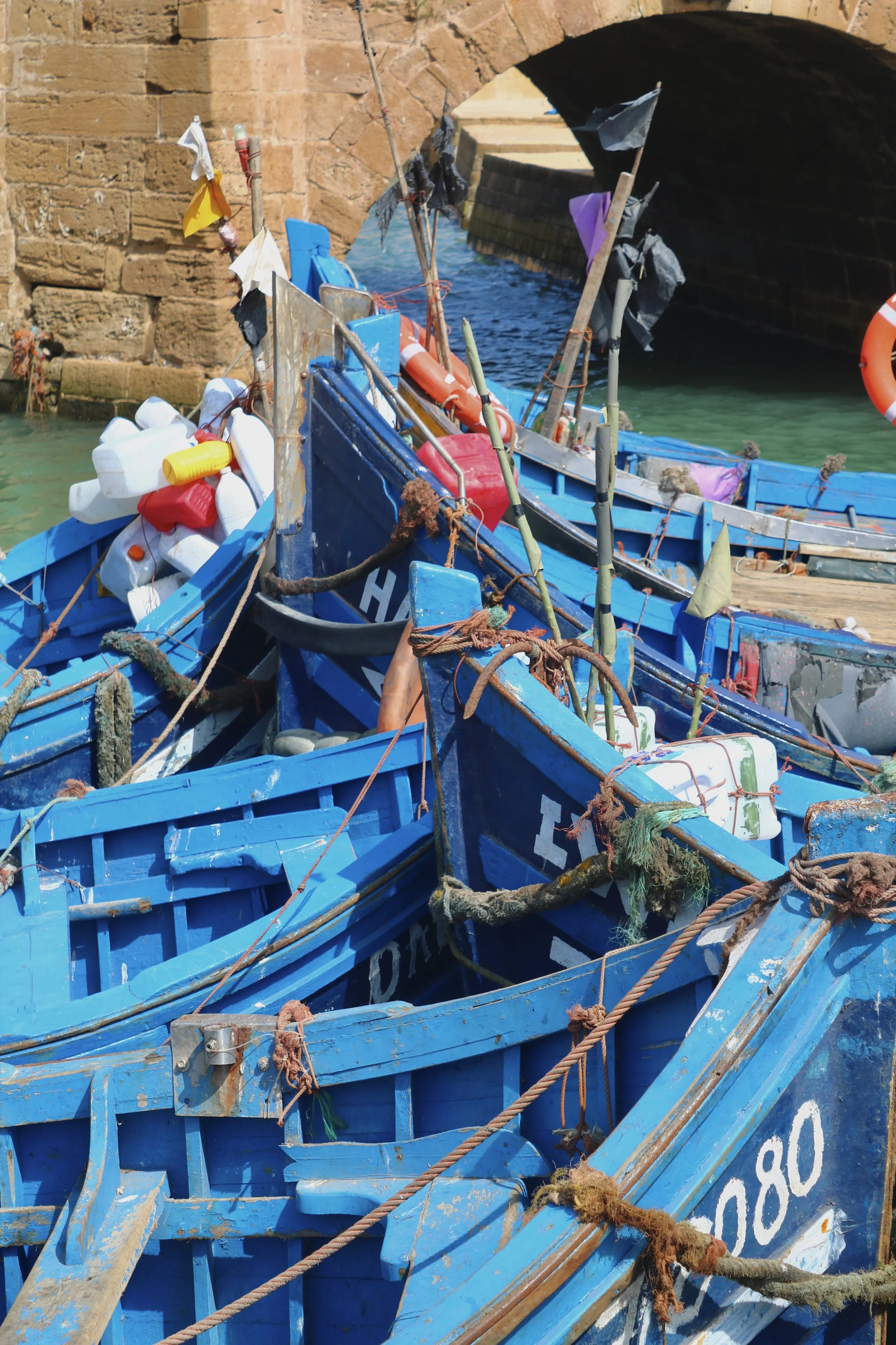 A collection of blue rowboats are tied up in the water near a stone arched bridge.