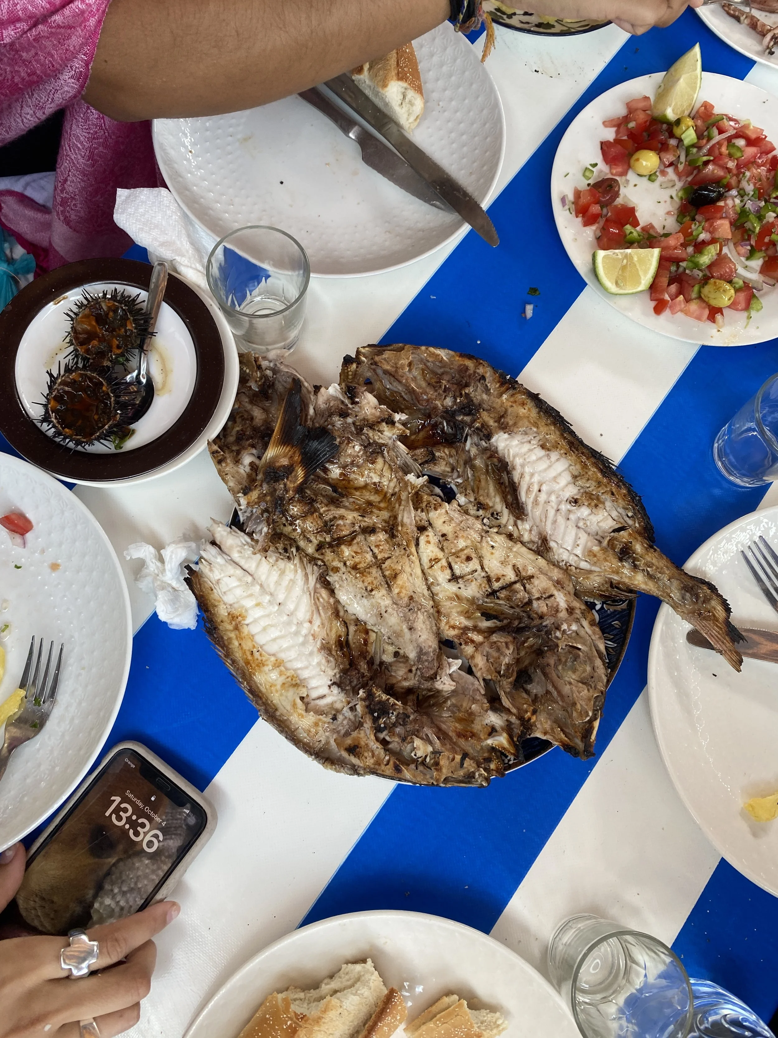 A plate of whole fish sits on a table with other plates with utensils and one plate has a salad.