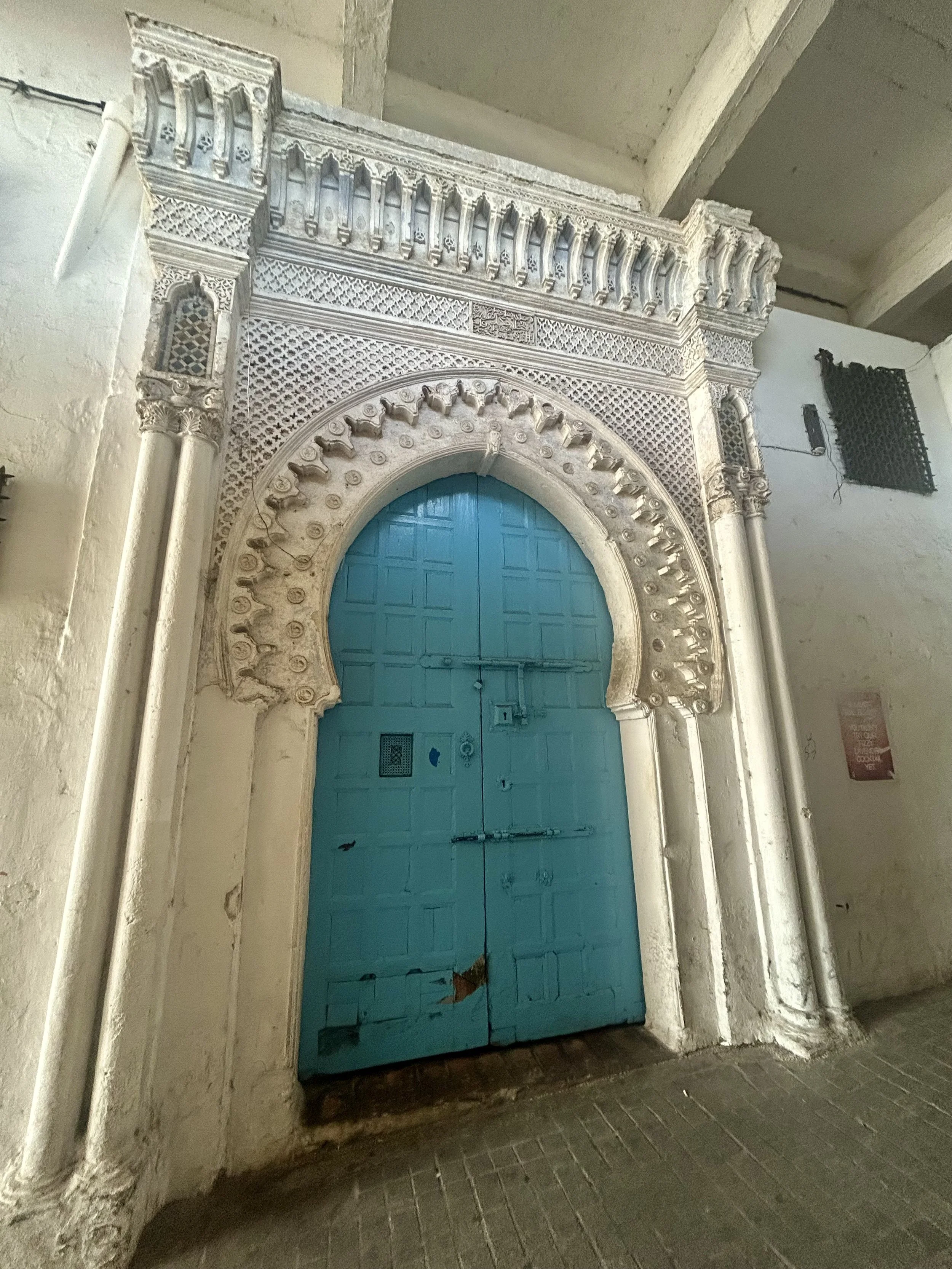 A bright blue door is framed by a white arch intricately decorated with a cut out pattern and arches on either side.