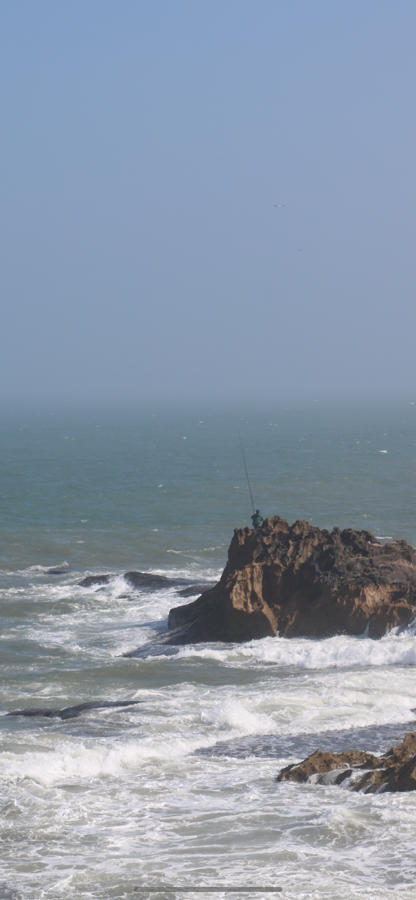 A fisherman fishes out on a rock that seems set in the middle of the ocean