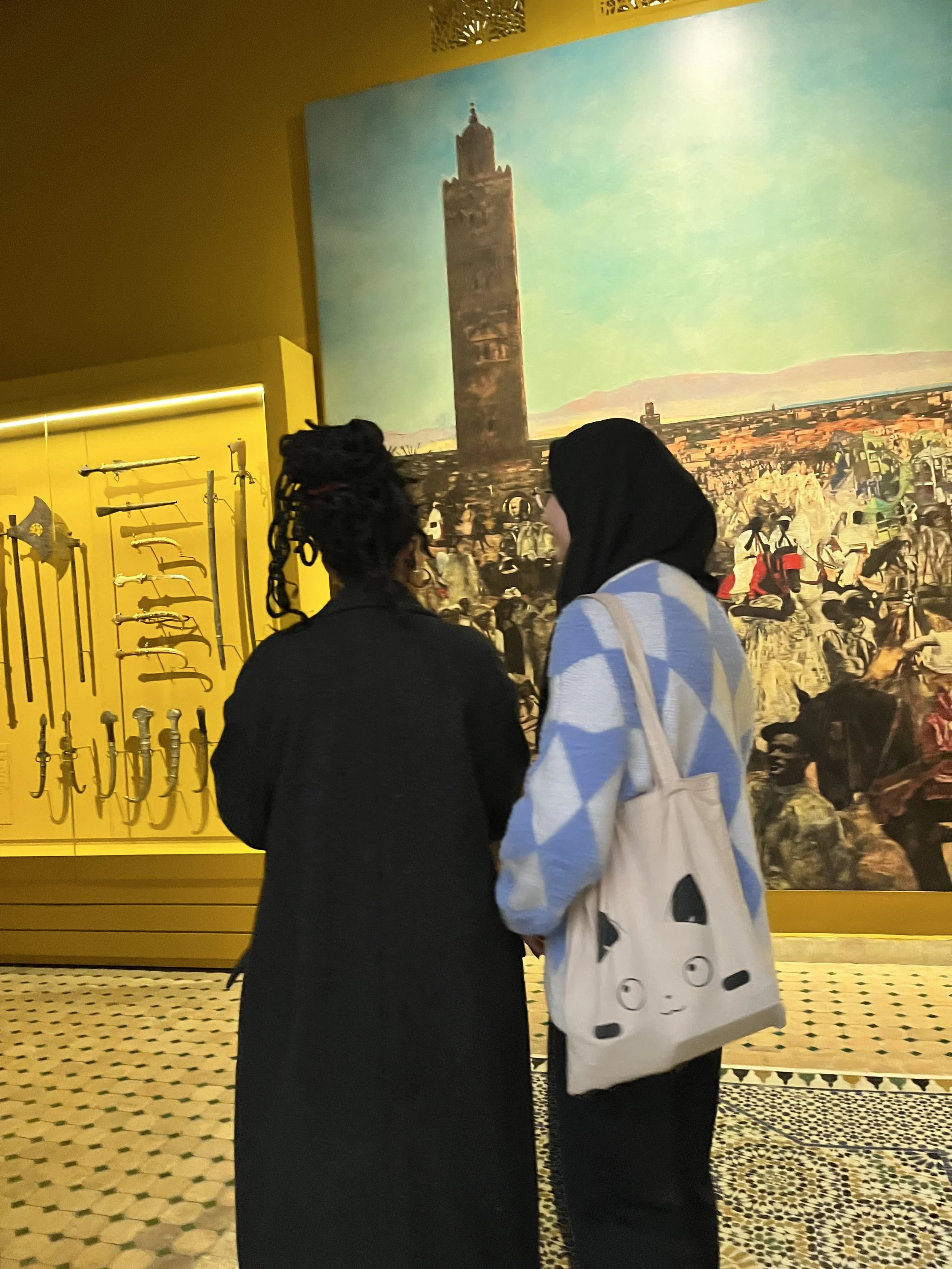 A photo from behind of two women looking at a large painting of a chaotic scene. To the left of the painting is a museum case with daggers and swords.