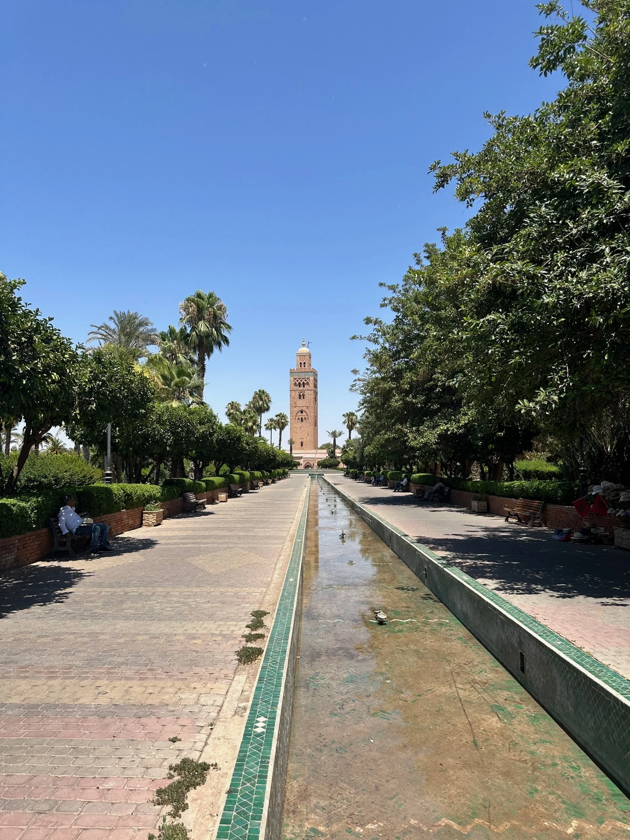 A long water trough lined by mosaic tiles and a patterned sidewalk leads up to the minaret of a mosque in the background. Trees line the path and people sit on benches in the shade on either side.
