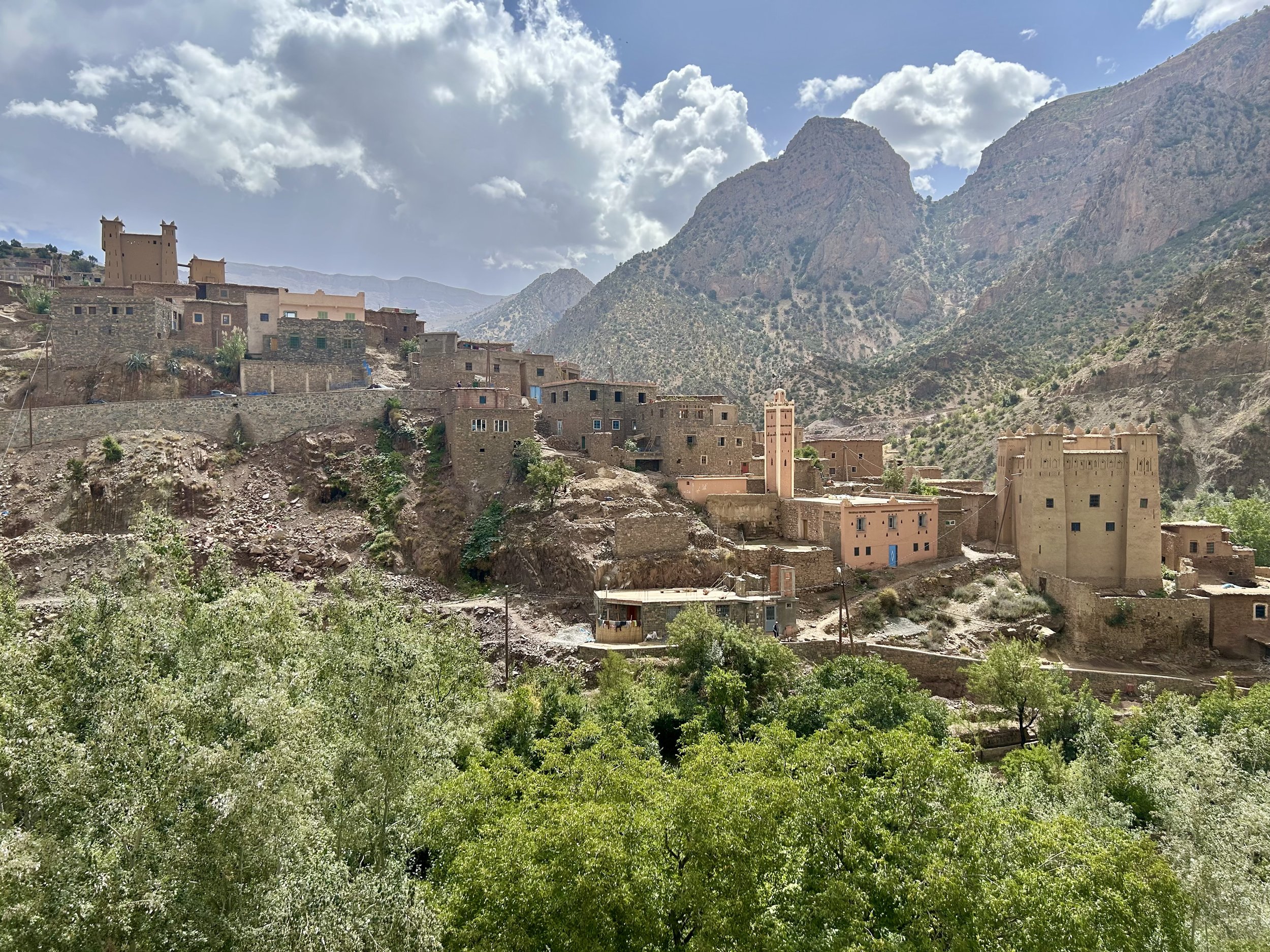 A town sits nestled among mountains and built from traditional red building materials. The mountains stretch up to the clouds.