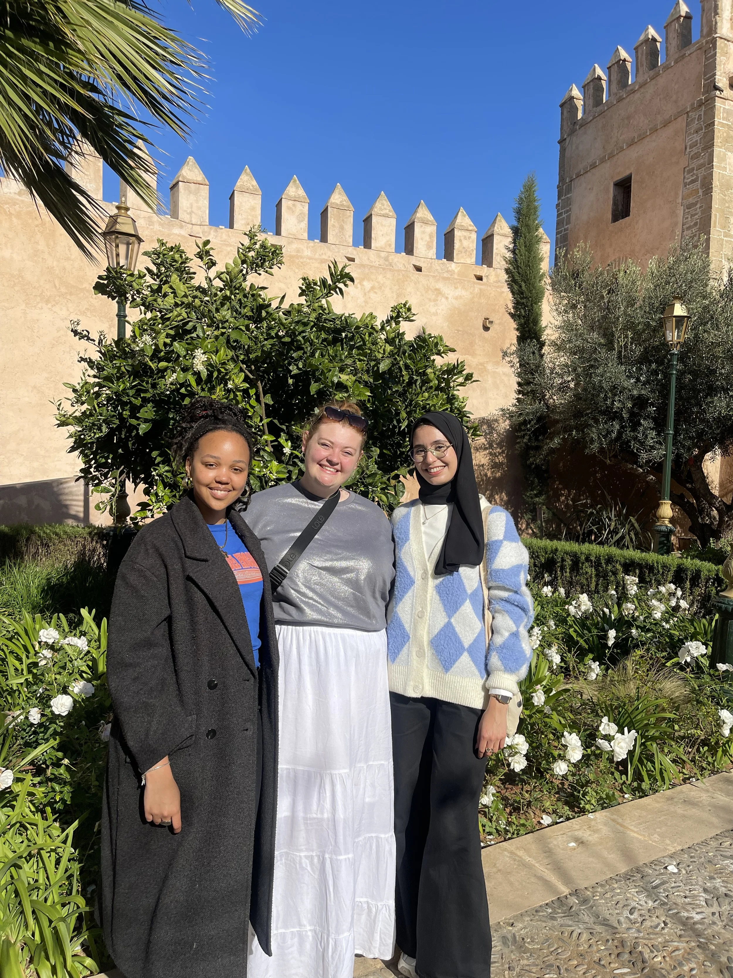 Three young women stand close together smiling in a sunny garden surrounded by high, castle-like walls.