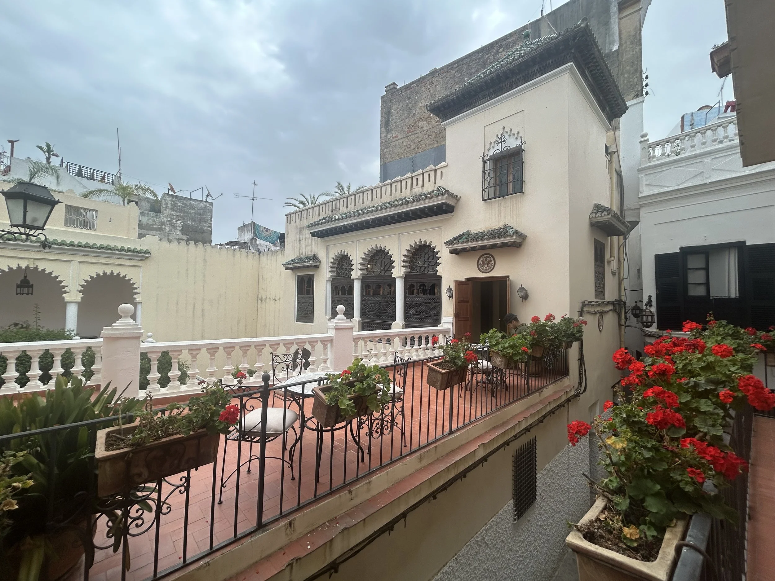 Taken from the second or third floor and looking onto a balcony of a white building with a courtyard. Geraniums sit in flower boxes