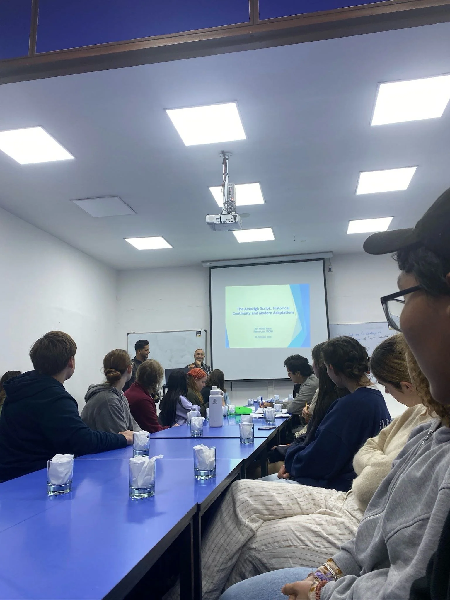 Students sit around a long table and listen to a teacher giving a lecture with a presentation at the front of the class.