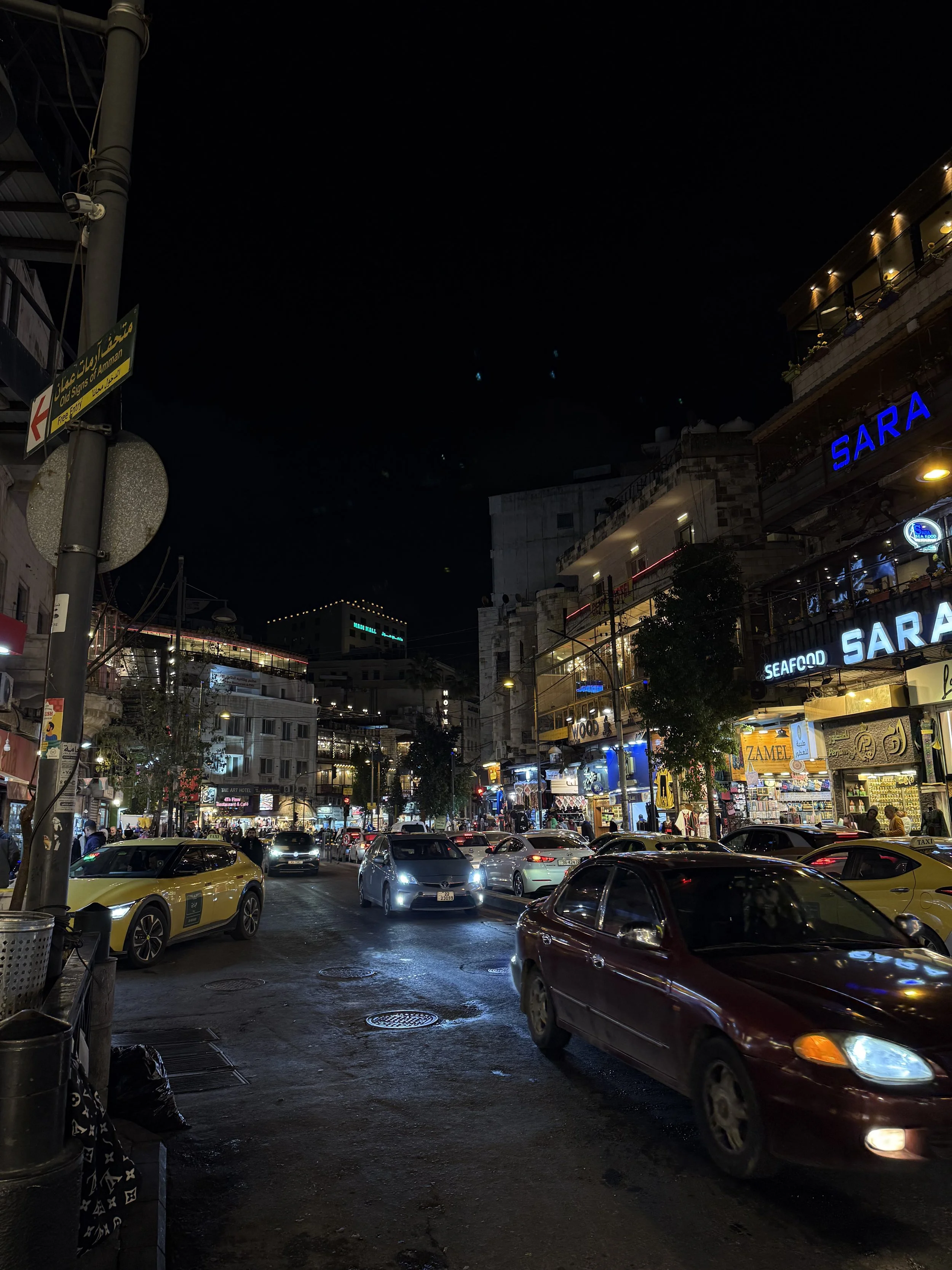 A busy night time street in Amman with shops, cars, and lots of people.