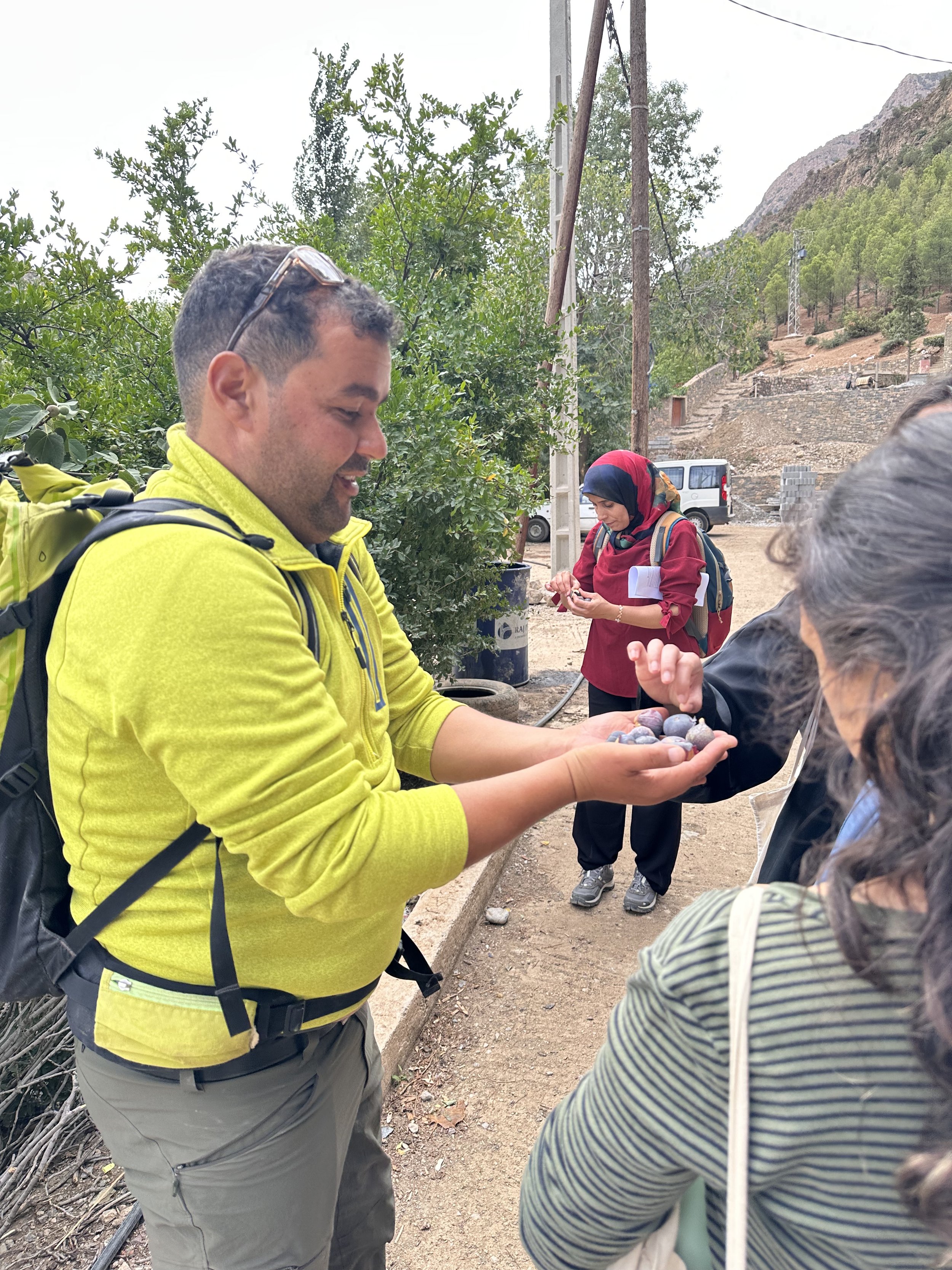 Along a dusty road lined with plants, an outdoor guide holds his hands full of small fruits out to a small group of people.