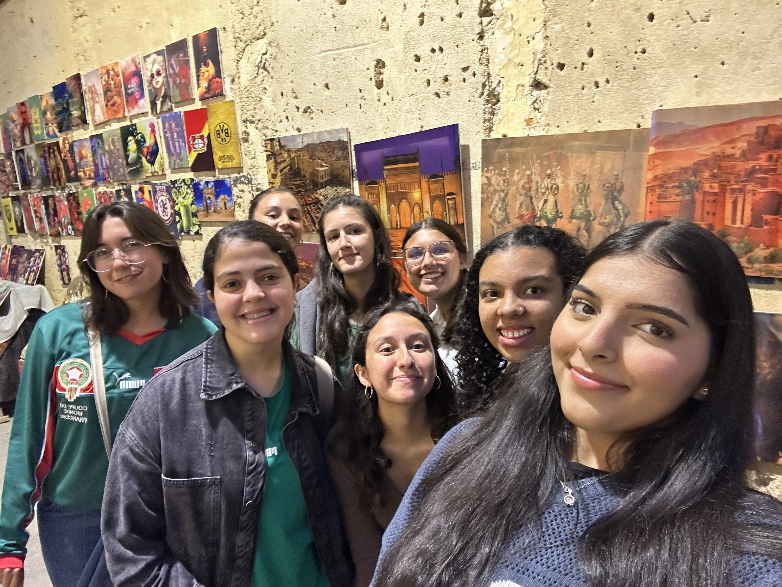 A group of 8 young women stand in a cluster smiling for the camera in front of a wall covered with paintings for sale.