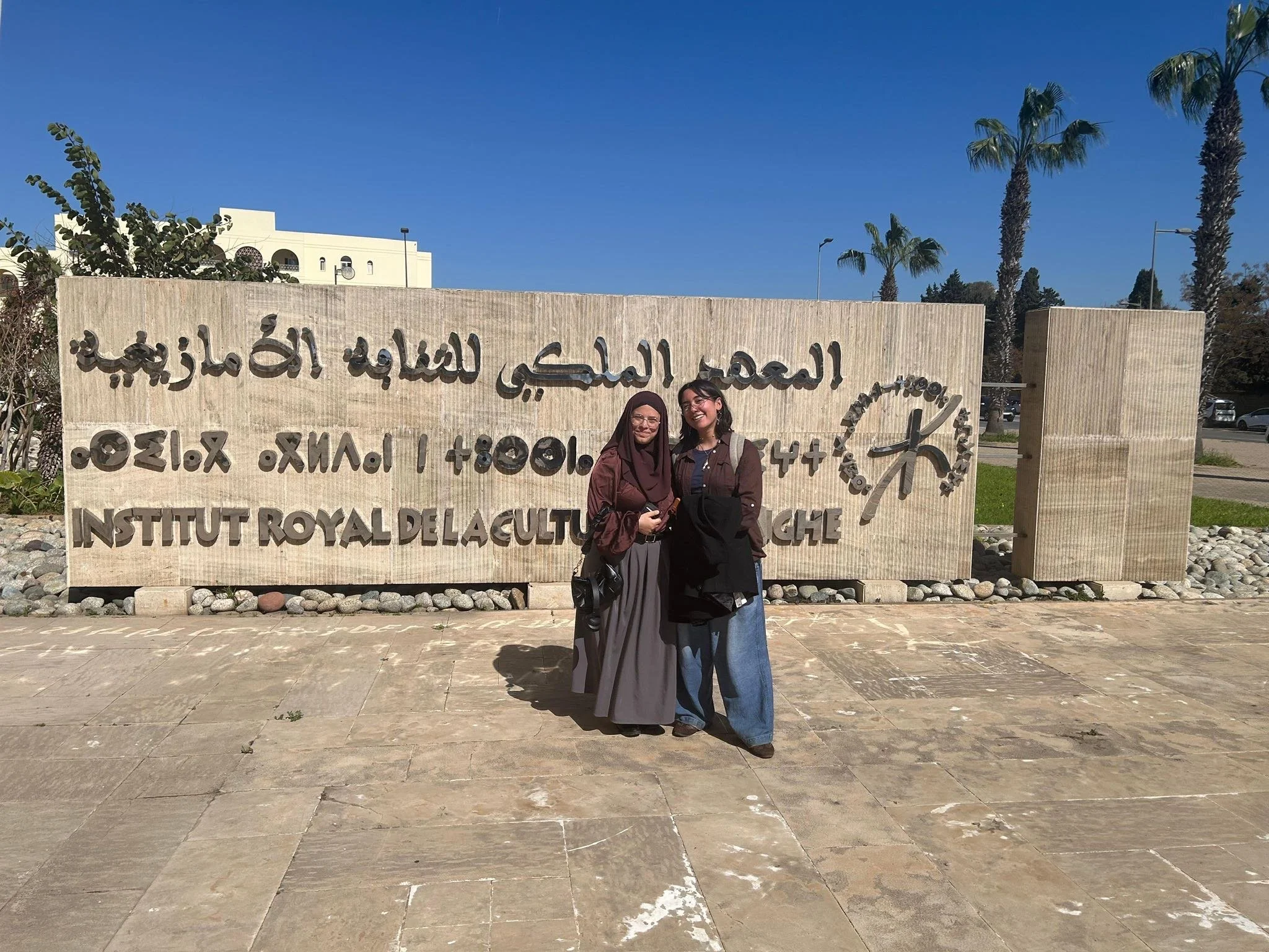 Nadia and a friend stand a smile outside a sign