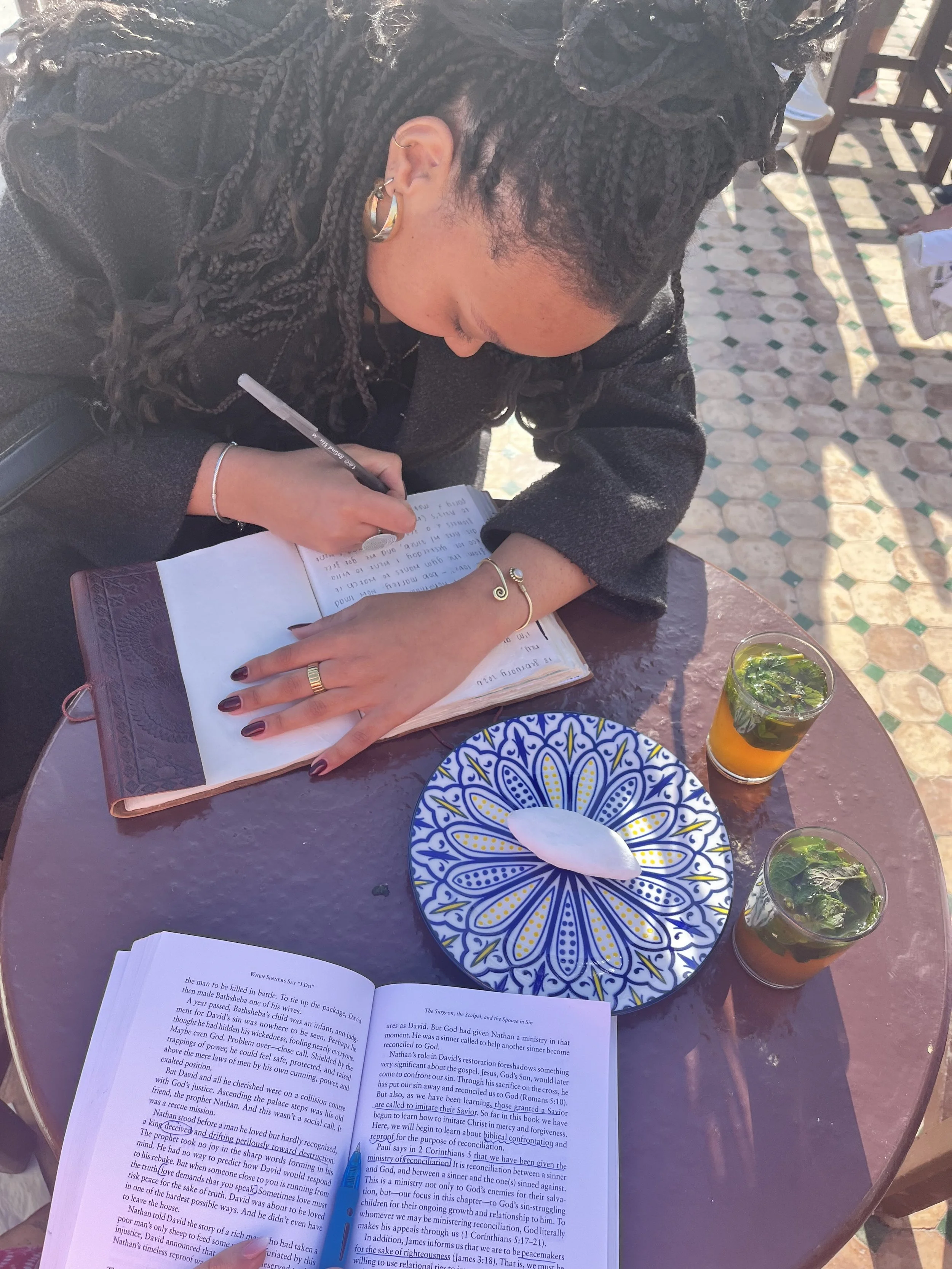 A young woman is seated at a cafe table, bent over as she writes in her journal