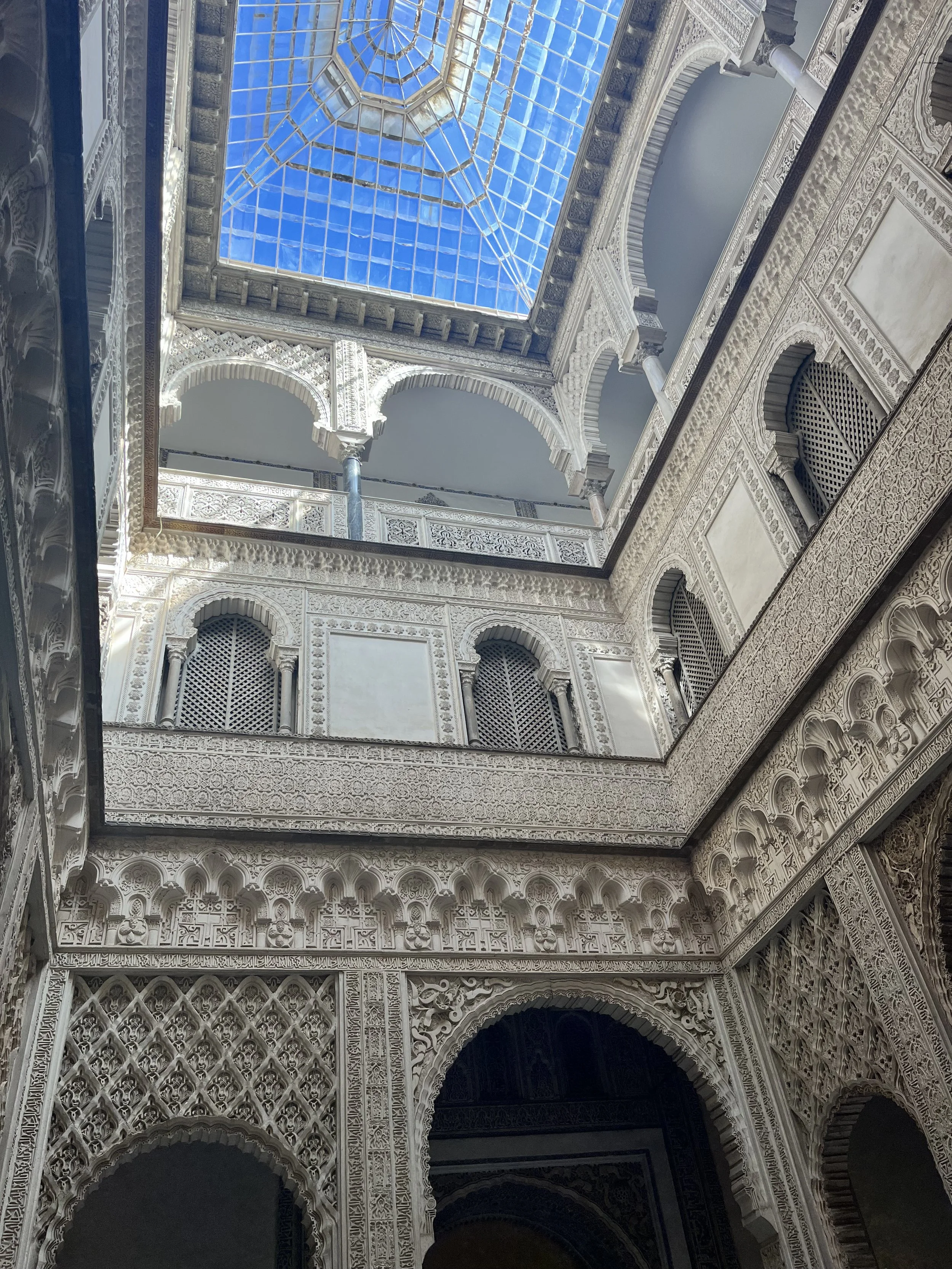 Looking up from the ground of an interior courtyard. The roof is made of small glass panes, and each story of the building is full of intricately carved windows and doorways in an Islamic style