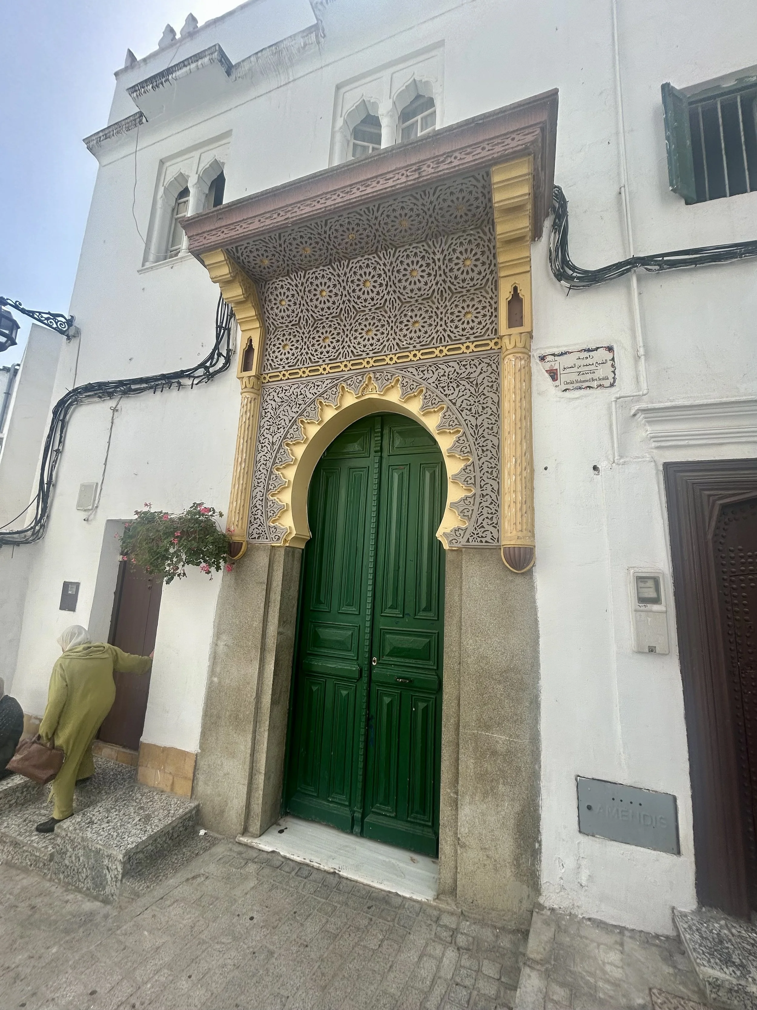 A large green door has an ornamental mosaic and wood cut arch and overhang over it. Set against a white wall background, the door stands out on the street.