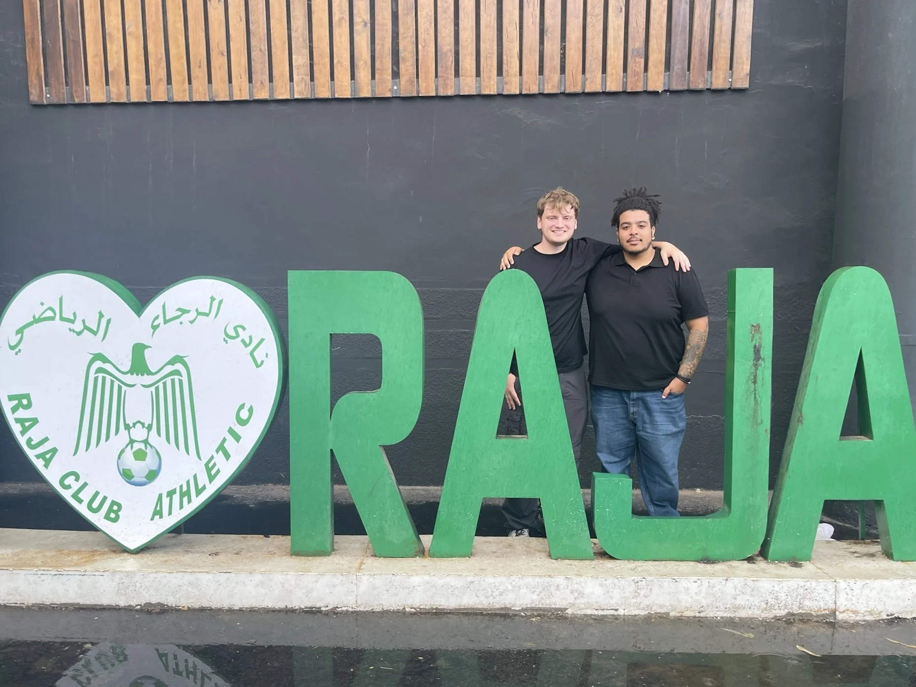Friends stand in matching black t-shirts behind a large sign that has a heart and then says "RAJA" in large letters