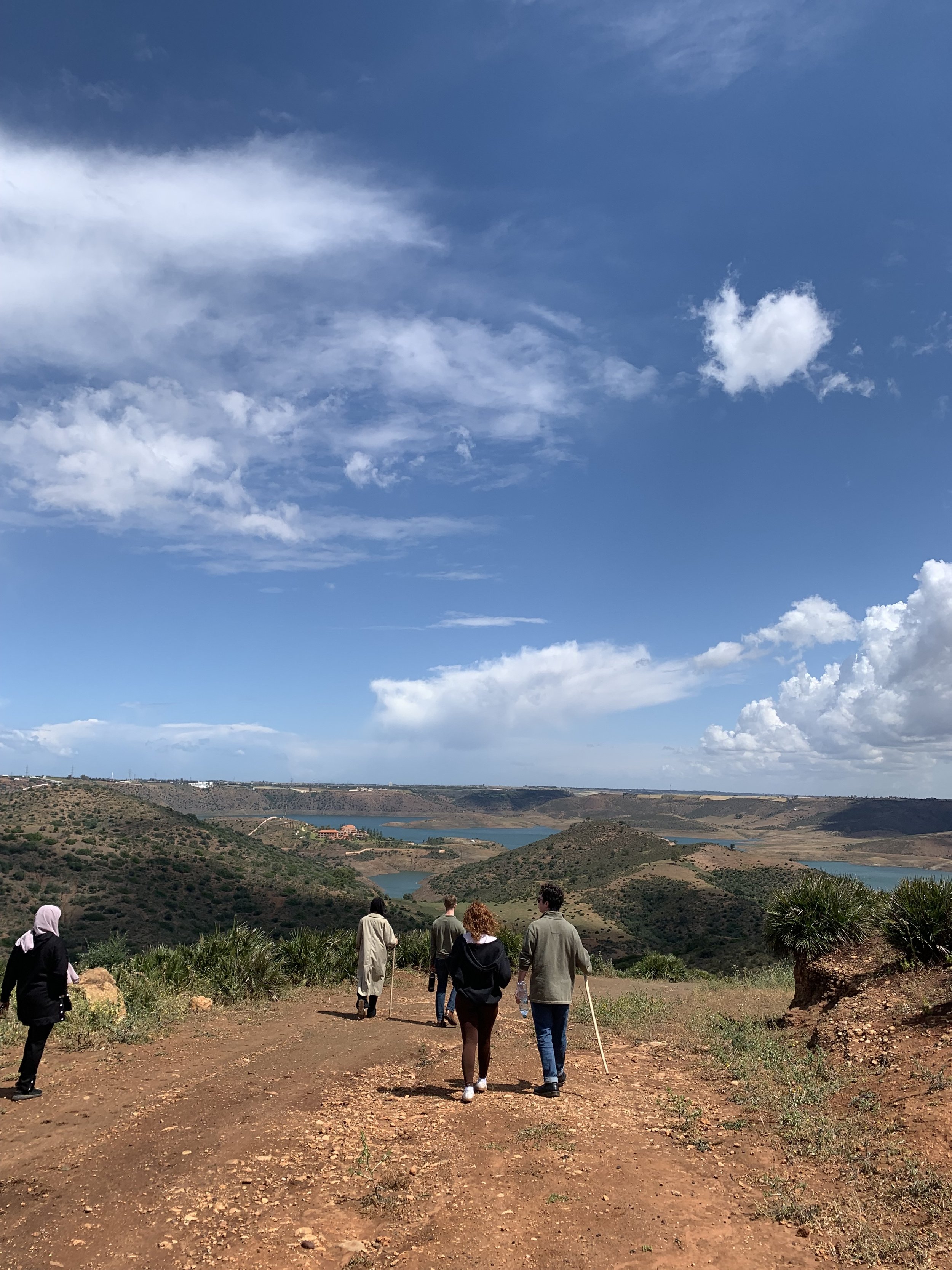 People walk on a large open trail on the mountains with small peaks and a river stretching out on the horizon.