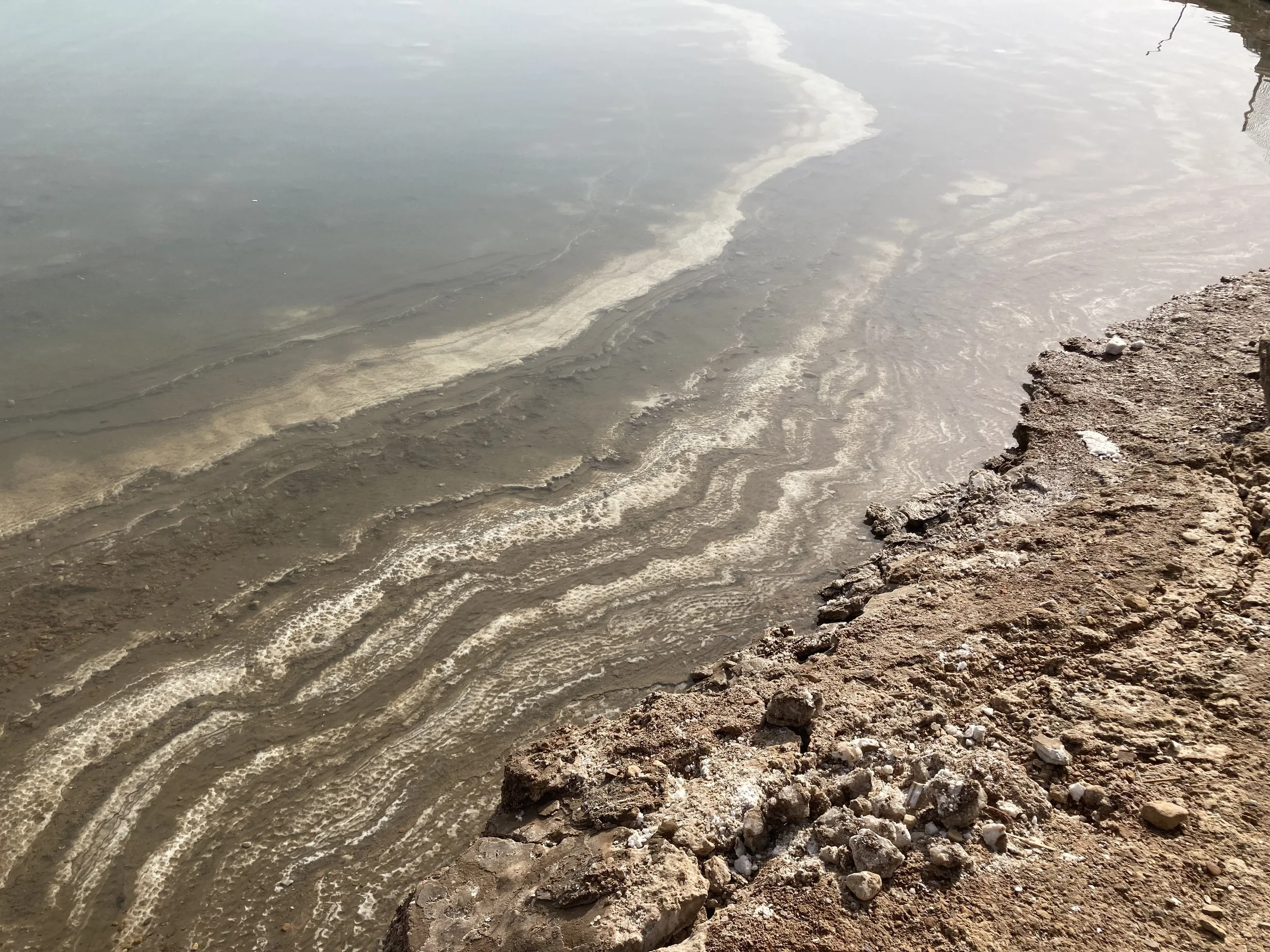 An aerial view of the shoreline shows a rocky beach, and white salt lines that show the receding water line.