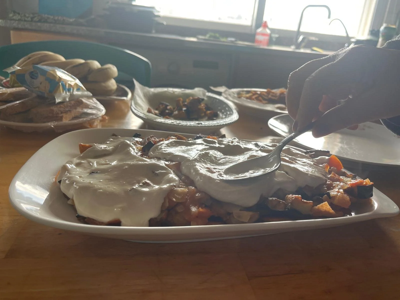 Someone spreads a creamy mixture on top of layers of eggplant and tomato in a kitchen