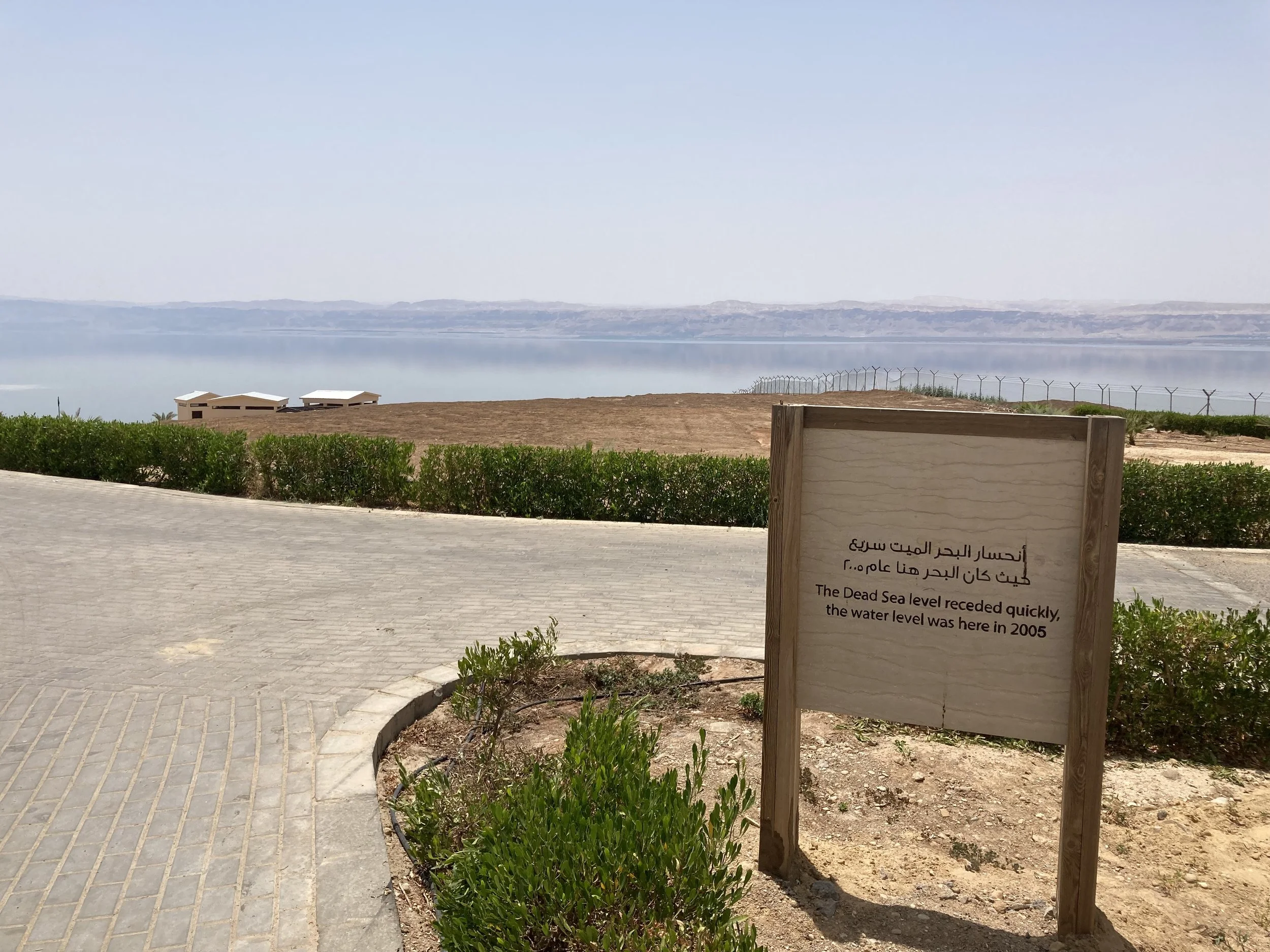An unassuming sign sits far from the water, along a paved path. It reads in English and Arabic "The Dead Sea level receded quickly, the water level was here in 2005."