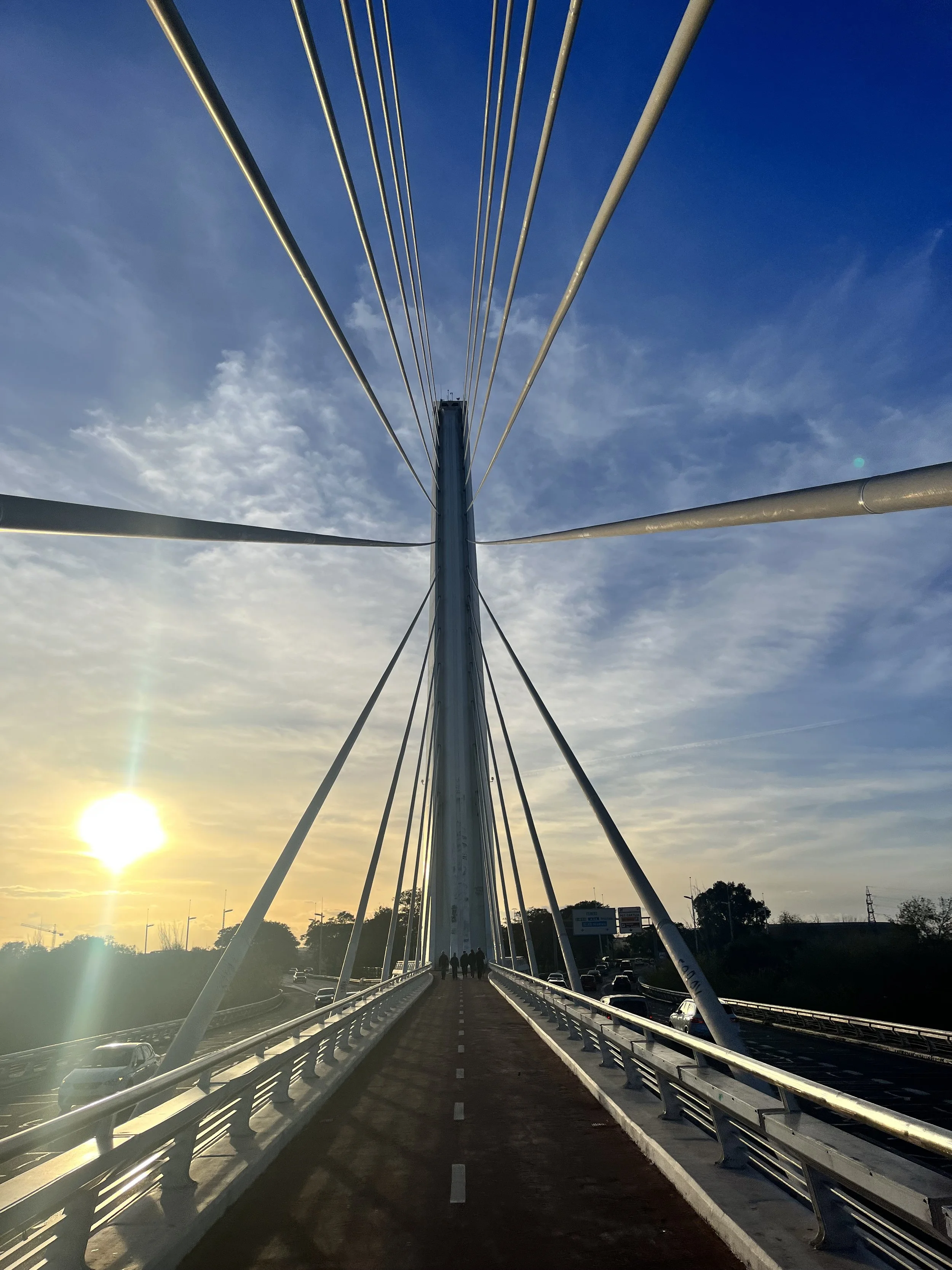 Looking down the pedestrian street across a narrow suspension bridge, framed on both sides by motor roads. The sun sits low on the horizon.