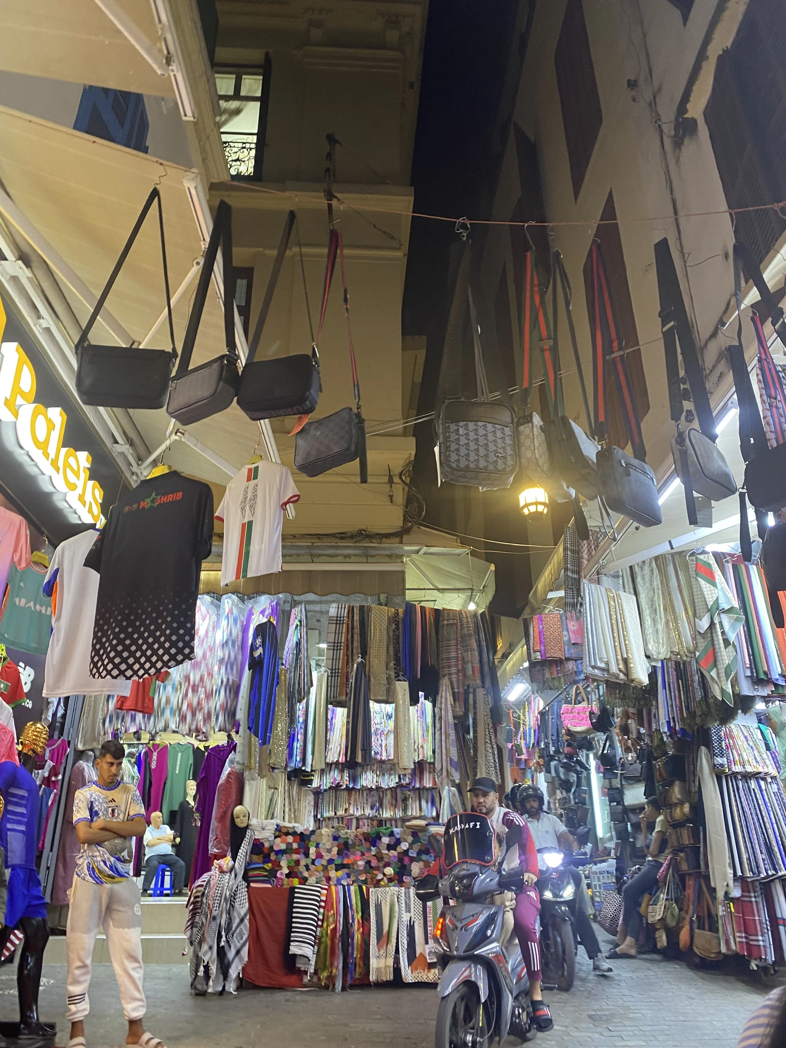 Taken at night, the market is extremely well lit. People drive their scooters through the narrow passages between shops selling all sorts of clothing, accessories, and textiles.