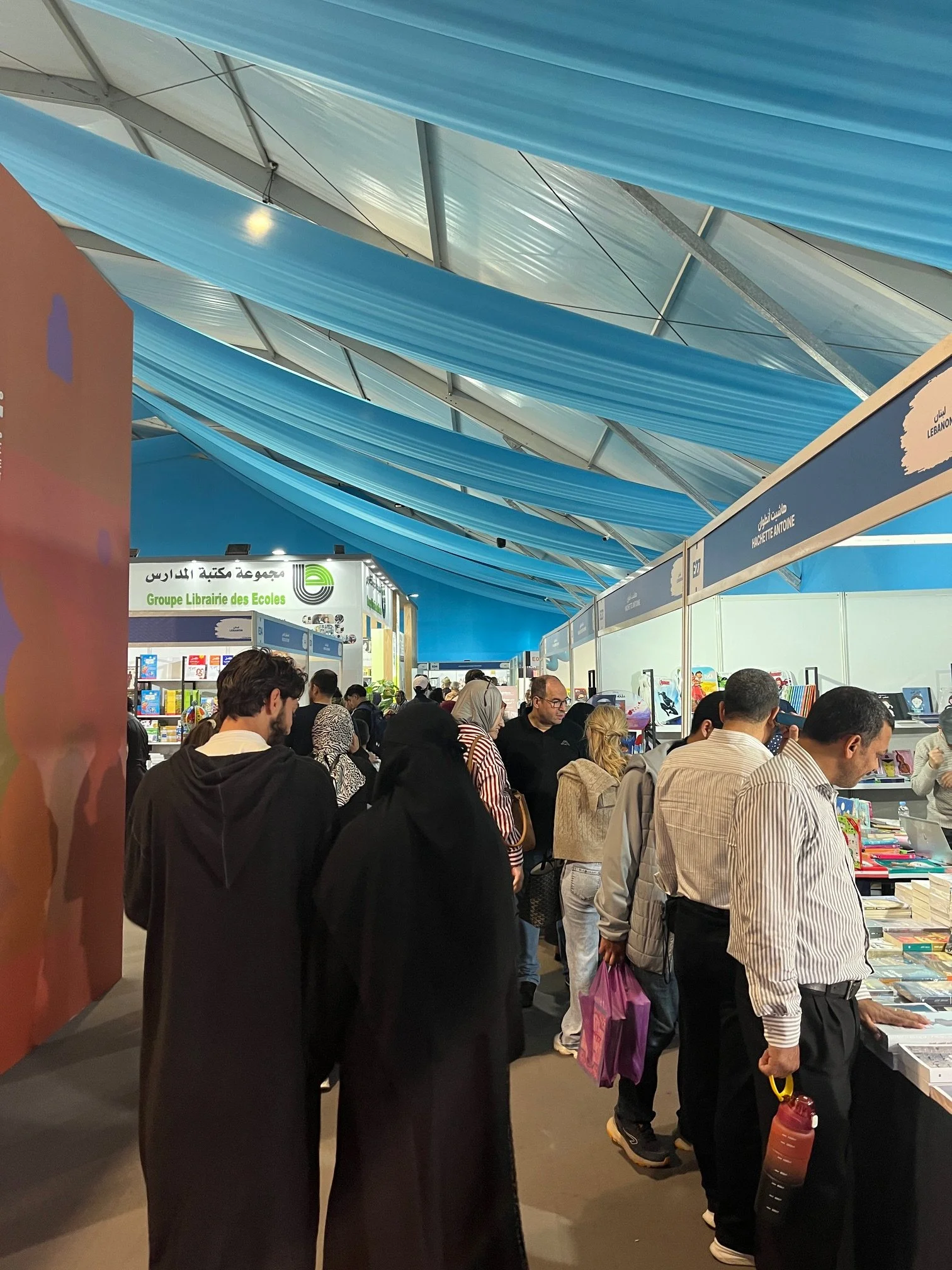 People walk in a crowded convention center type space looking at stalls of books.