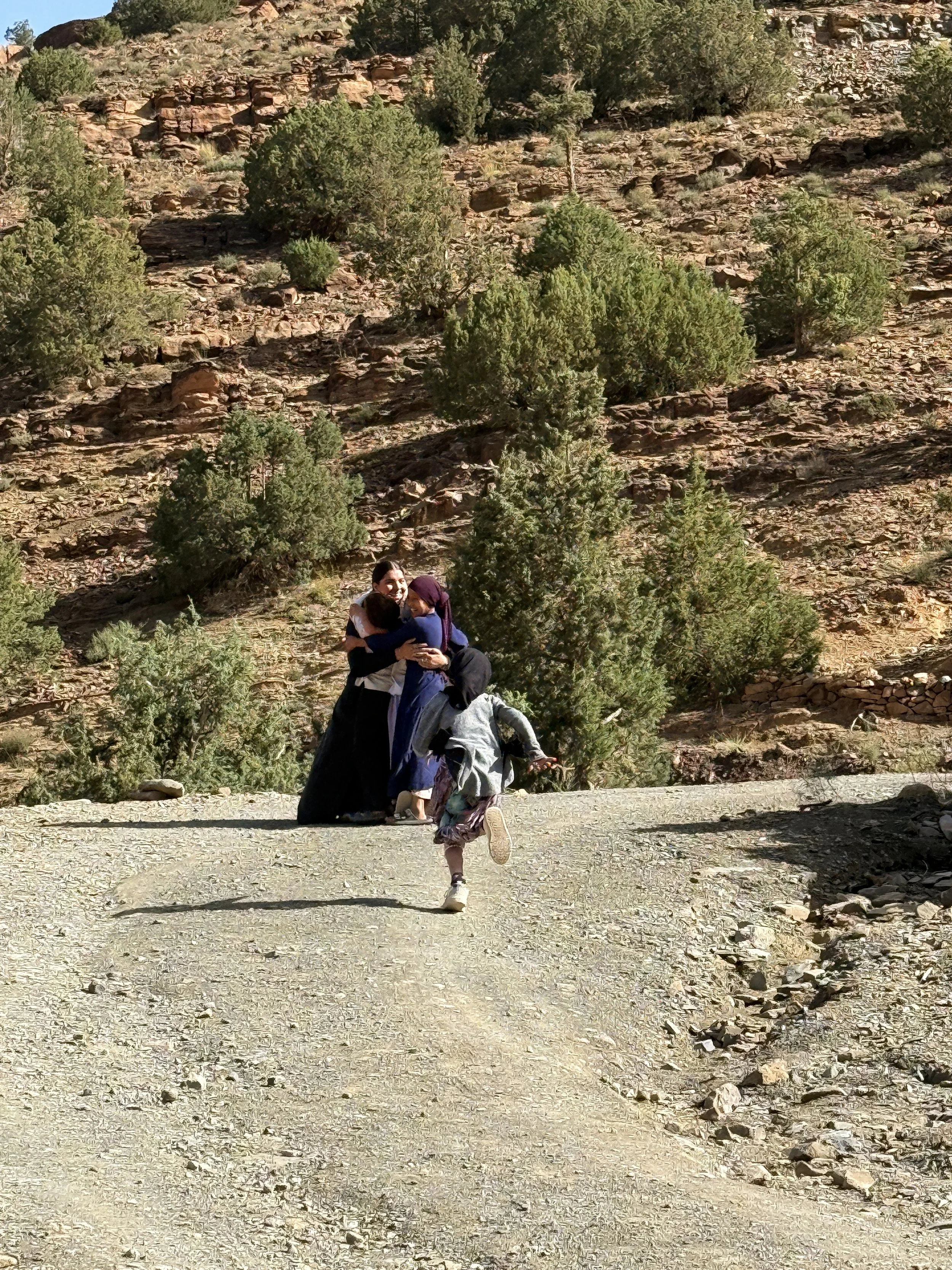 Outside on a gravel road in shrubby mountains, four young people are hugging.