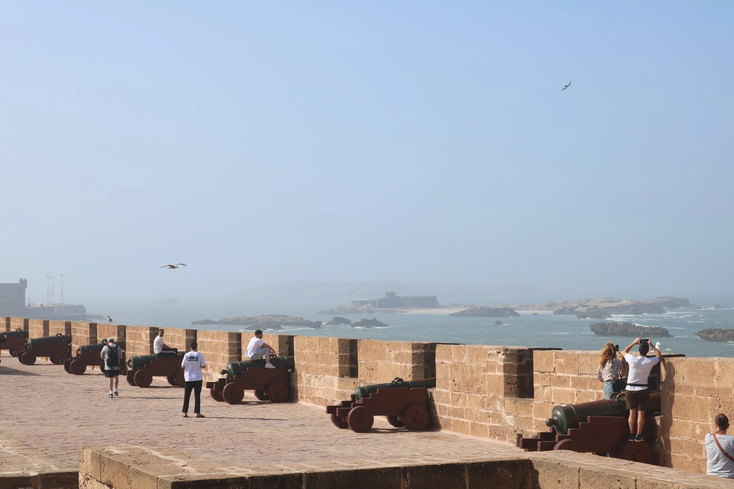 On top of the wall of a fortress built on the sea with cannons pointed out towards the water and tourists walking around