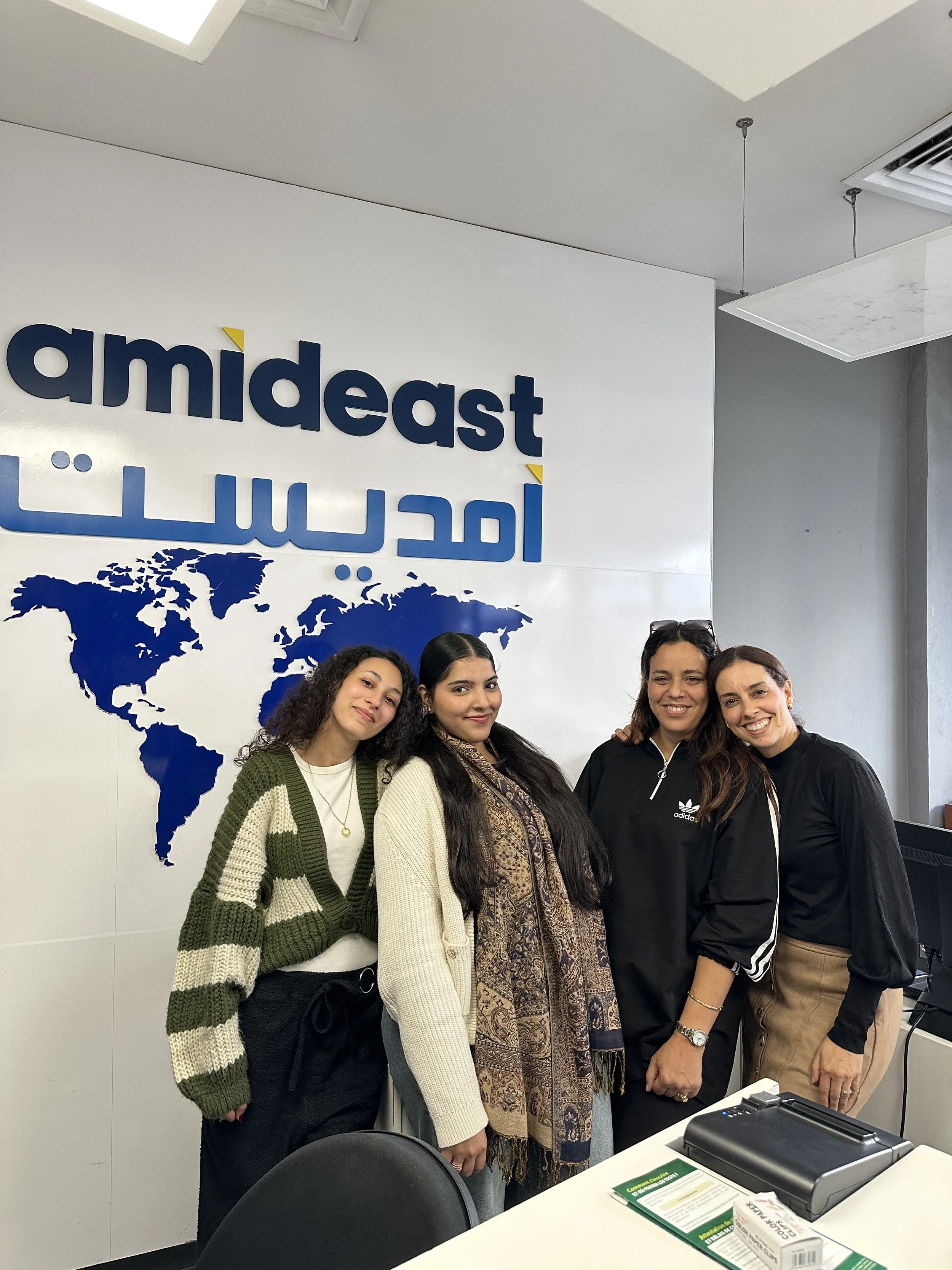 Four young women stand side by side smiling for the camera behind a desk in front of an Amideast sign.