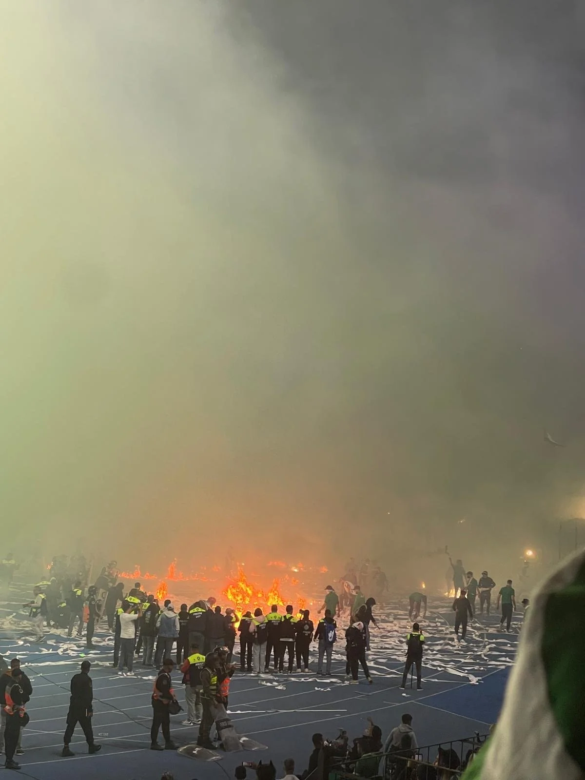 A fire on a running track with a large circle of emergency responders watching on and a sky filled with smoke
