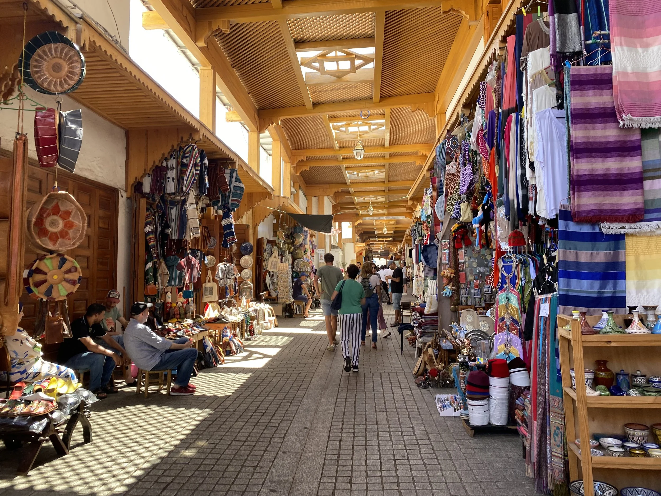 Looking down the market alley between shops selling textiles, shoes, poofs, and more. The market is covered by carved wooden panels.