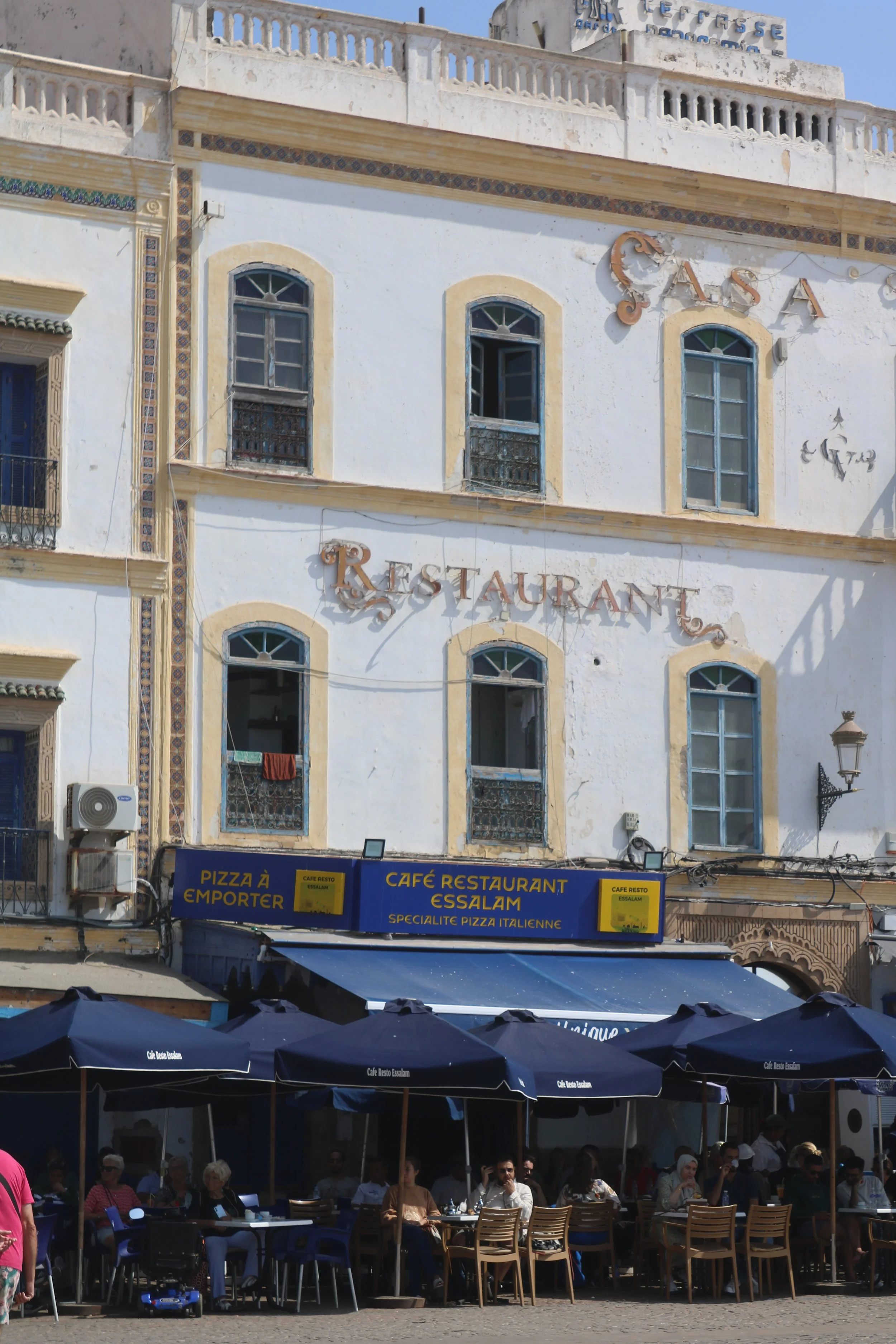 People sit at tables at a cafe under blue umbrellas at a building that has several stories and is white and appears old.
