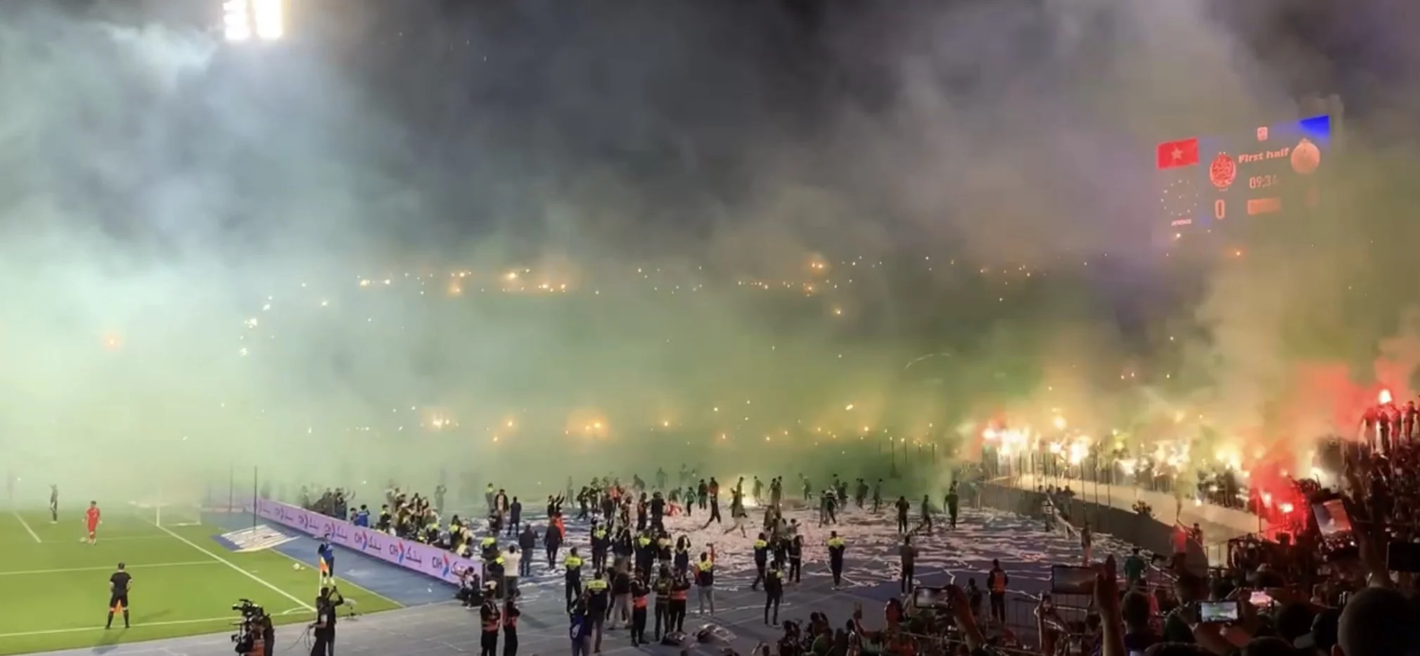 Green, red, and black smoke comes from another end of the football pitch
