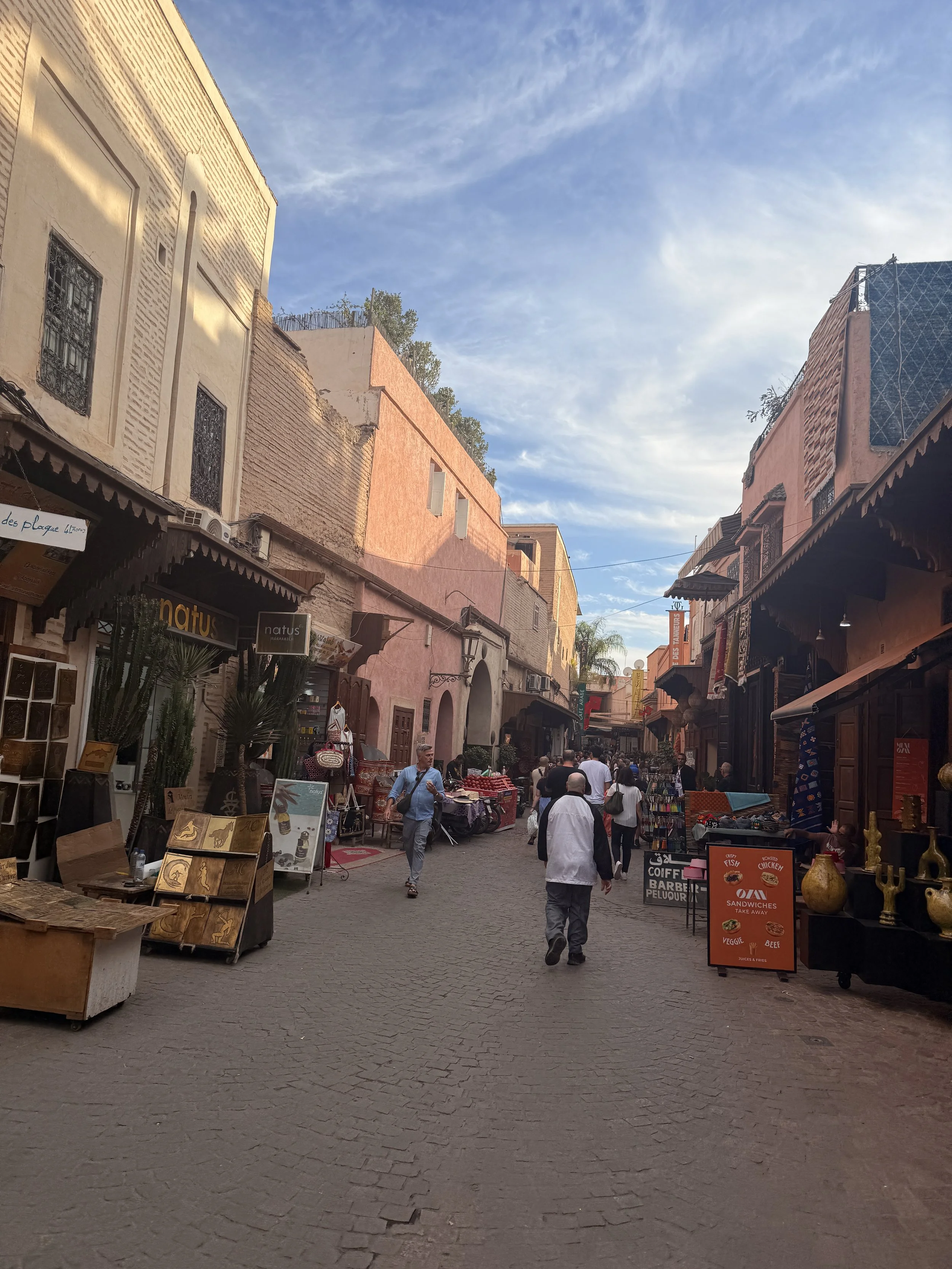 A quiet market street with few people. Goods are set out in front of shops and signs advertise coffee and snacks.