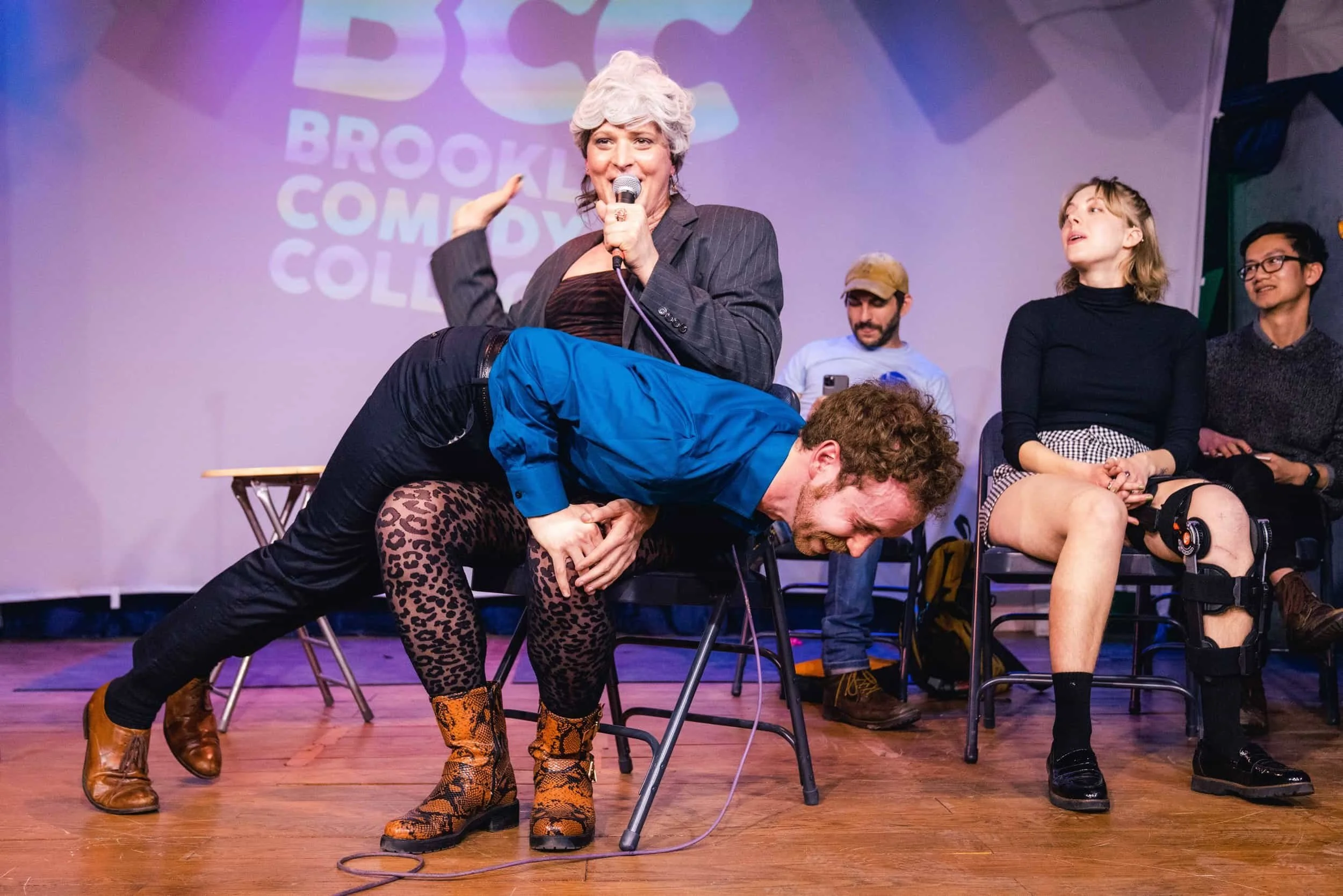 Photo of performers on stage that predominantly features one sitting in a chair with a white wig, with another laying across their lap