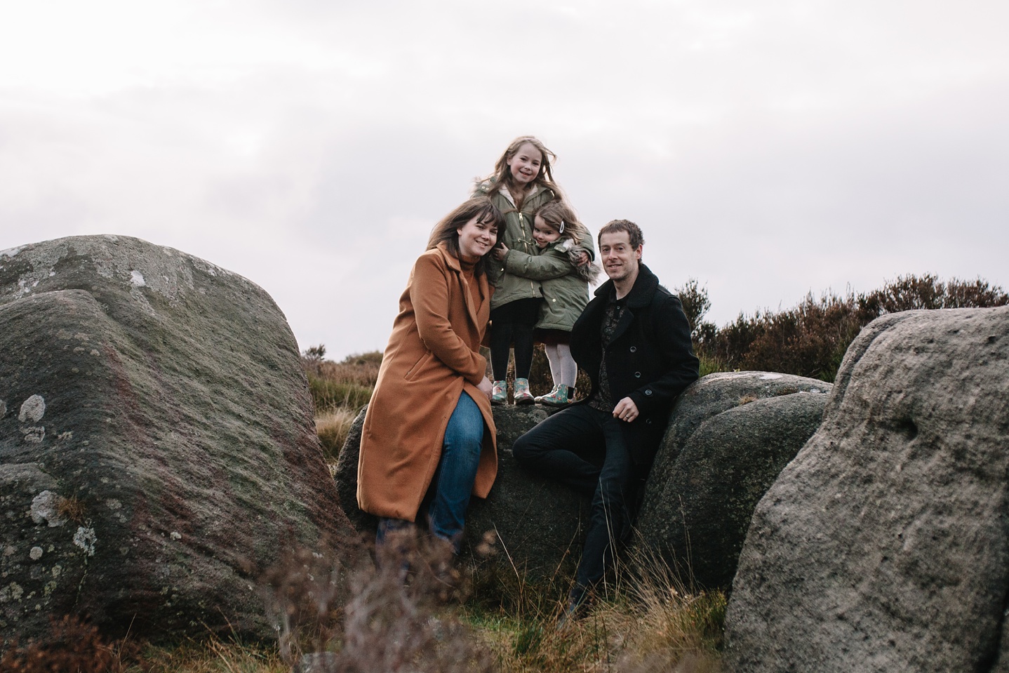 mum and dad sat on a rock with two little girls stood up beside them smiling at the camera