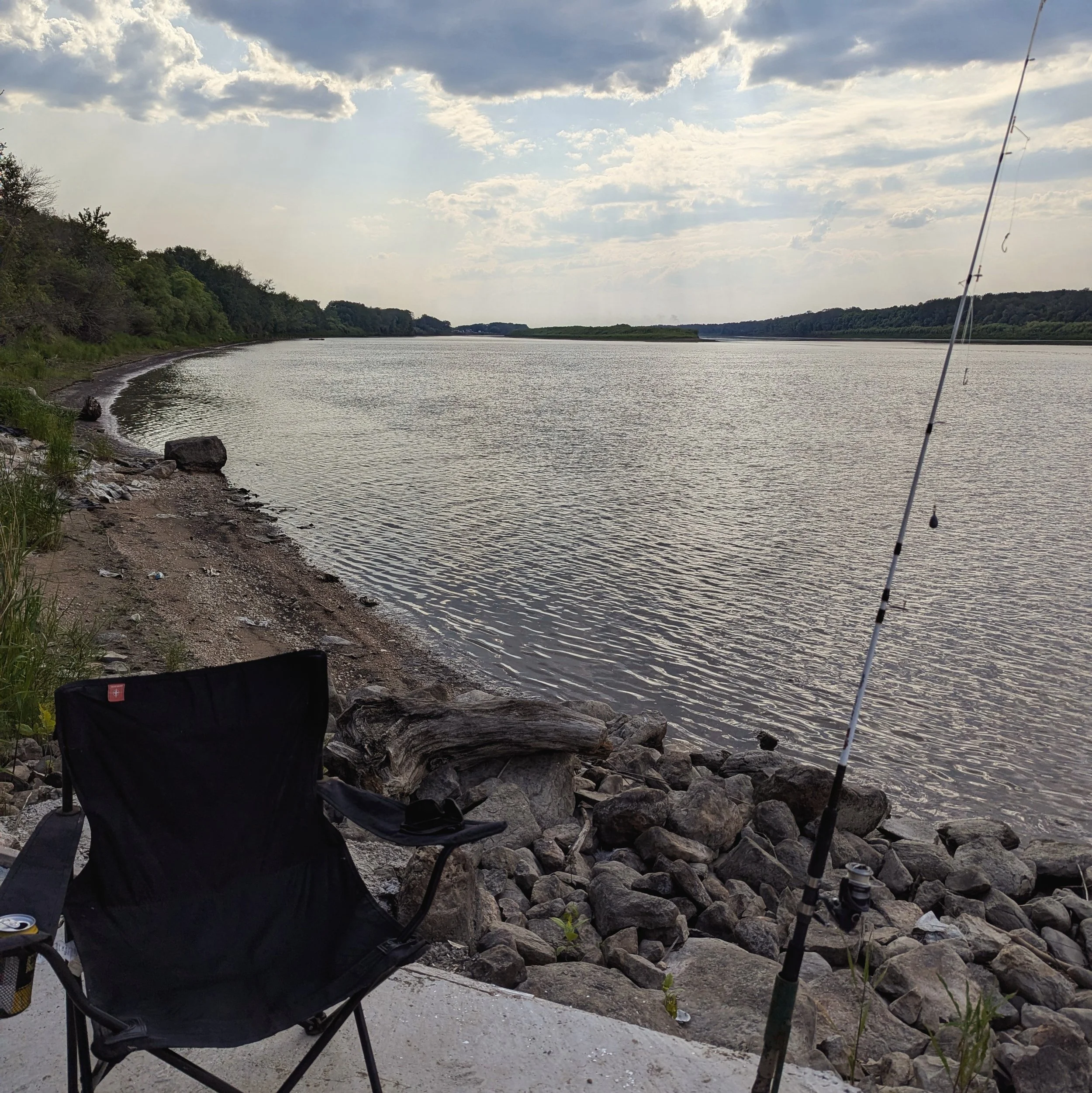 A fishing rod set up on rocky shore beside a black camping chair, overlooking a calm river with trees on the horizon and a partly cloudy sky.