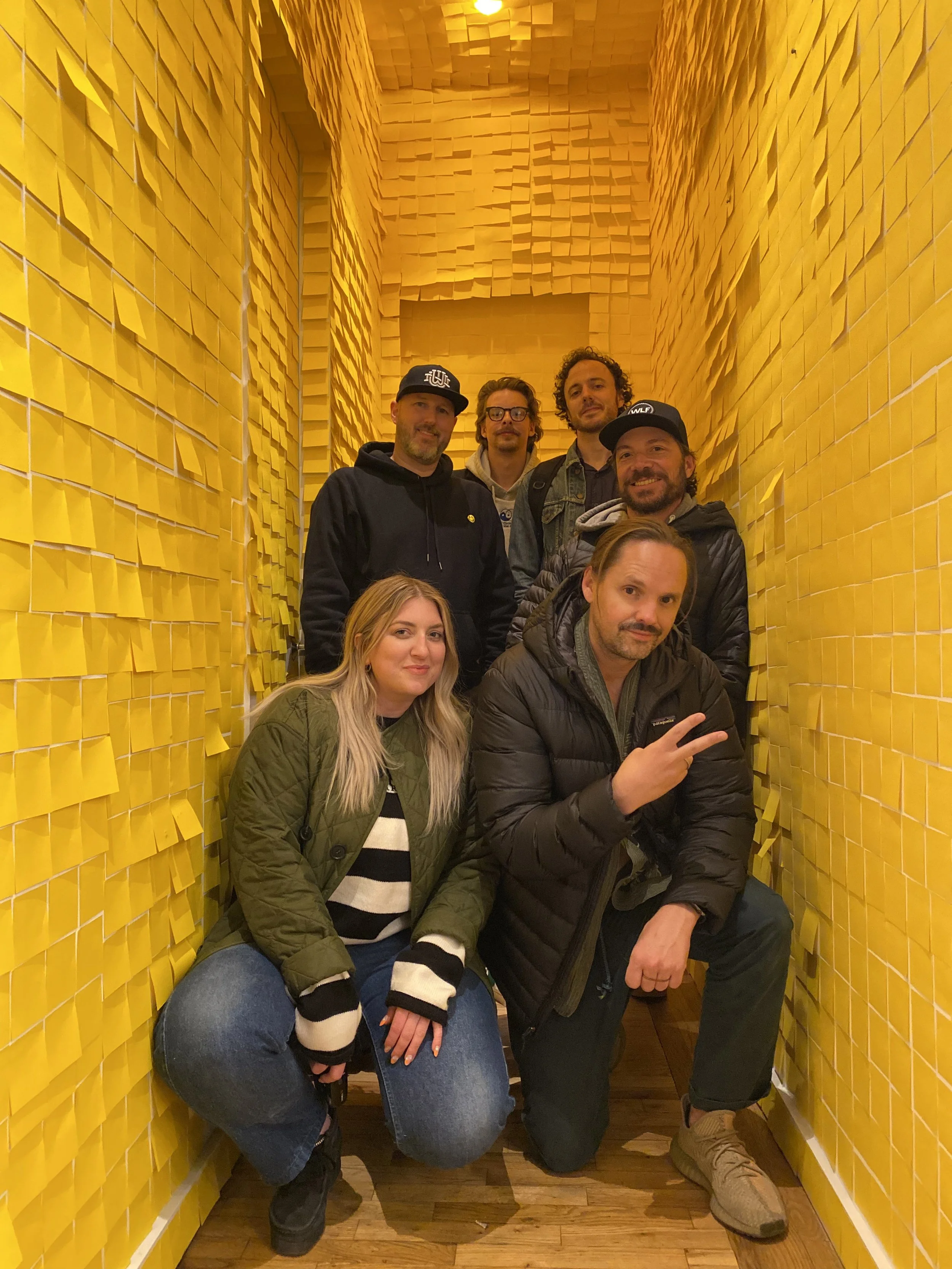 Seven people posing in a small, yellow, tiled corridor with sticky notes on the walls.