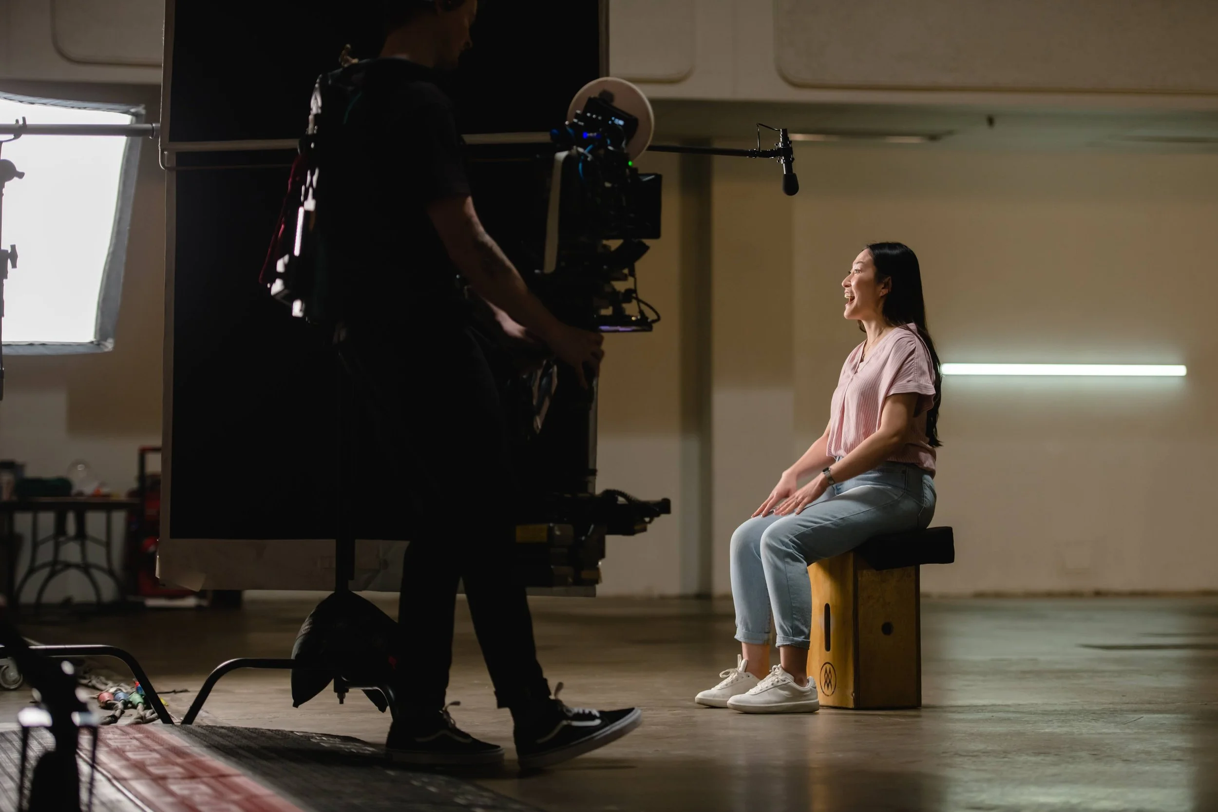A woman with long dark hair wearing a pink top and light blue jeans sitting on a wooden box and talking during a video shoot in a spacious indoor studio, with a cameraman filming her.