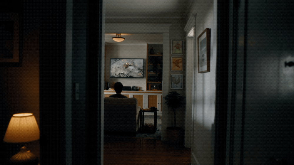 A person sitting on a sofa in a living room, watching television, as seen from a hallway with a table lamp on the left and wall art on the right.